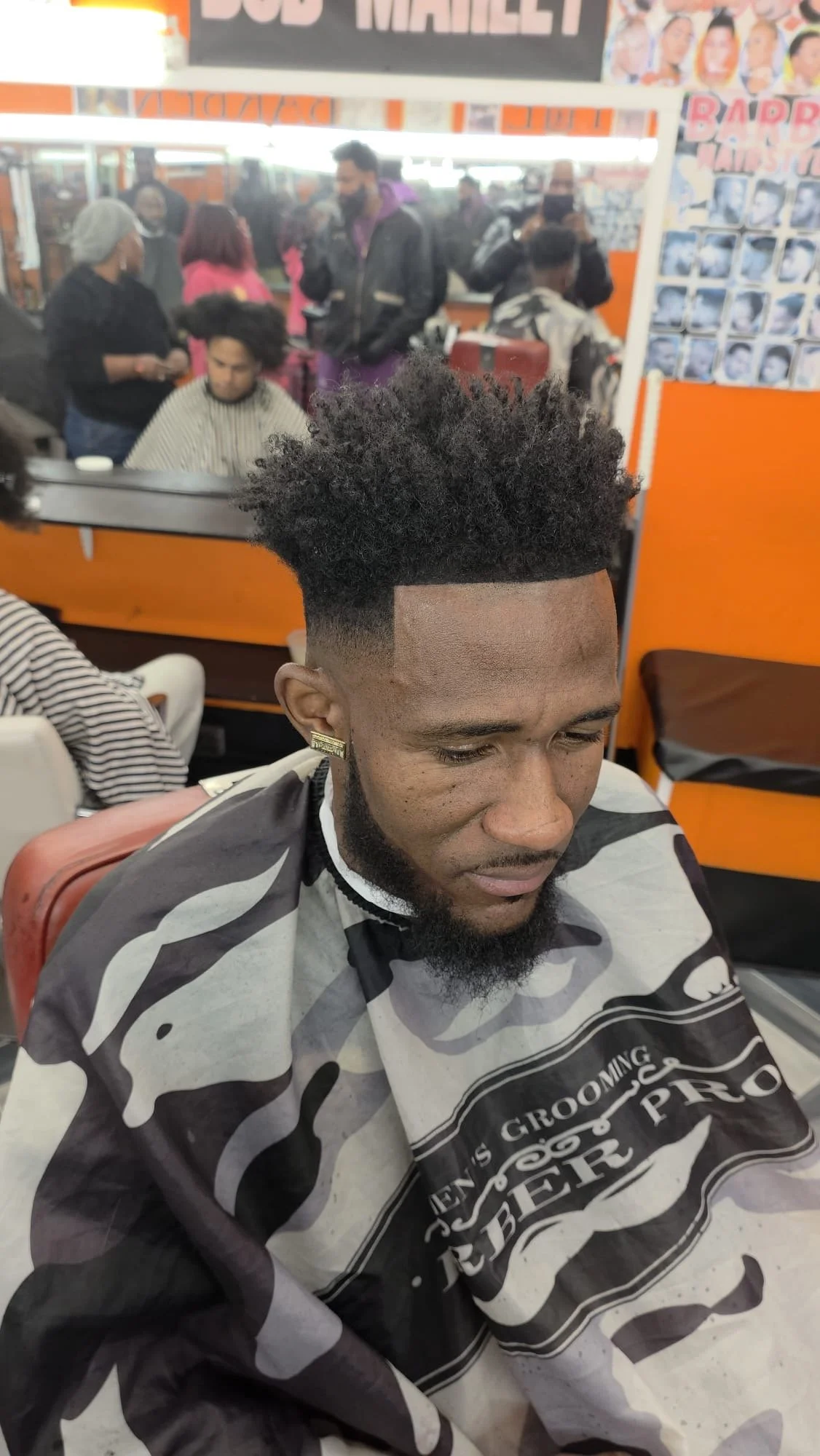 A man with a fresh high top fade haircut in a barber shop, wearing a barber cape with camouflage pattern. Behind him are other clients and barbers, some with capes, inside the salon.