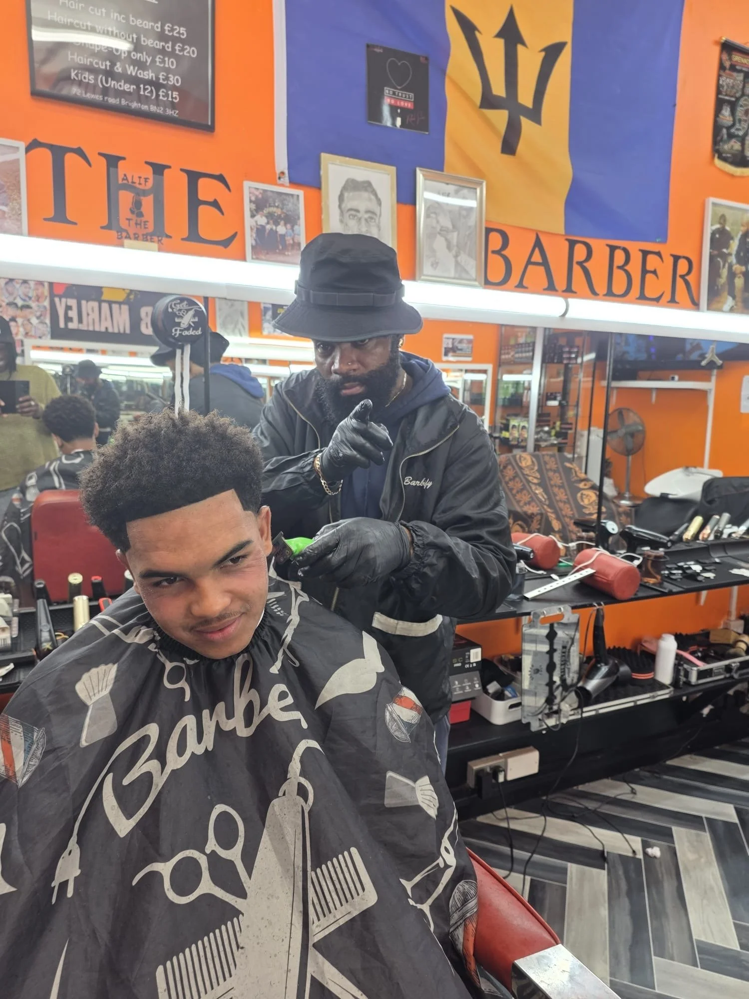 A man getting a haircut at a barber shop, with a barber working on his hair. The barber is pointing upward with his finger and wearing black gloves. The customer is sitting in a chair with a cape featuring barber-themed graphics. The background has orange walls, a mirror, and various barber tools and prints, including a flag on the wall.