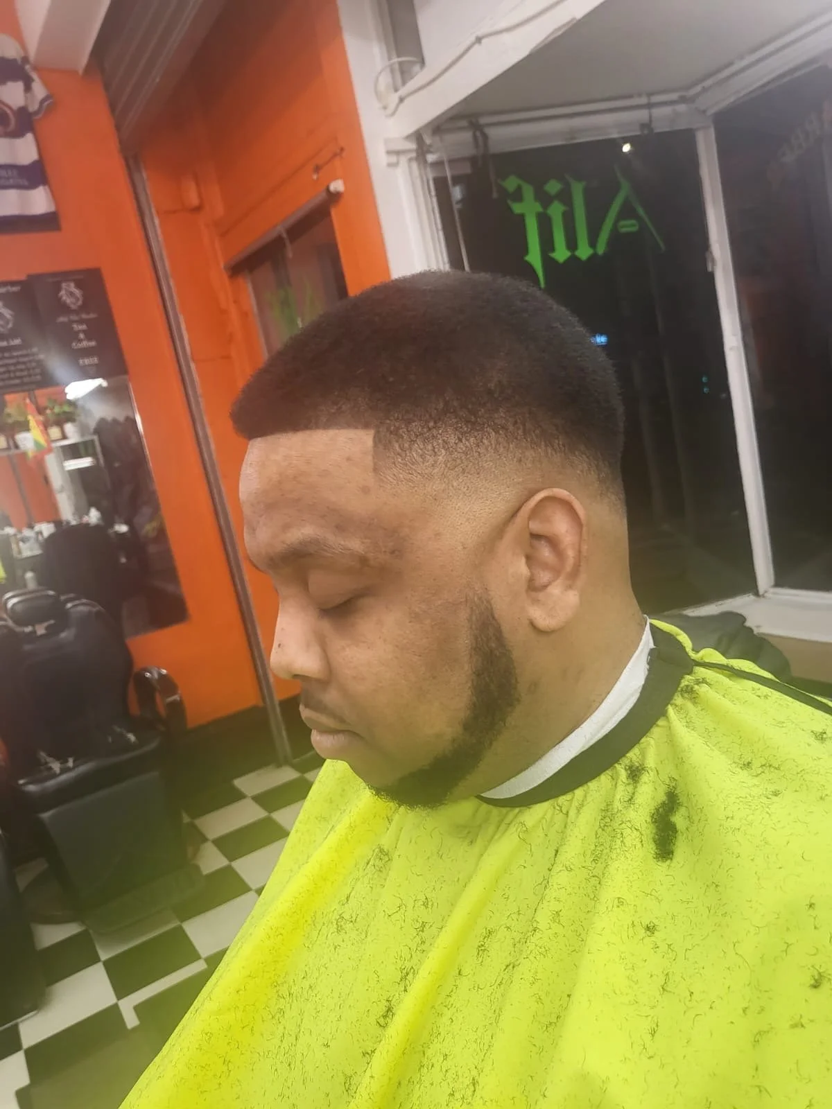 Young man with a fresh fade haircut sitting in a barber shop with a yellow barber cape draped over him.