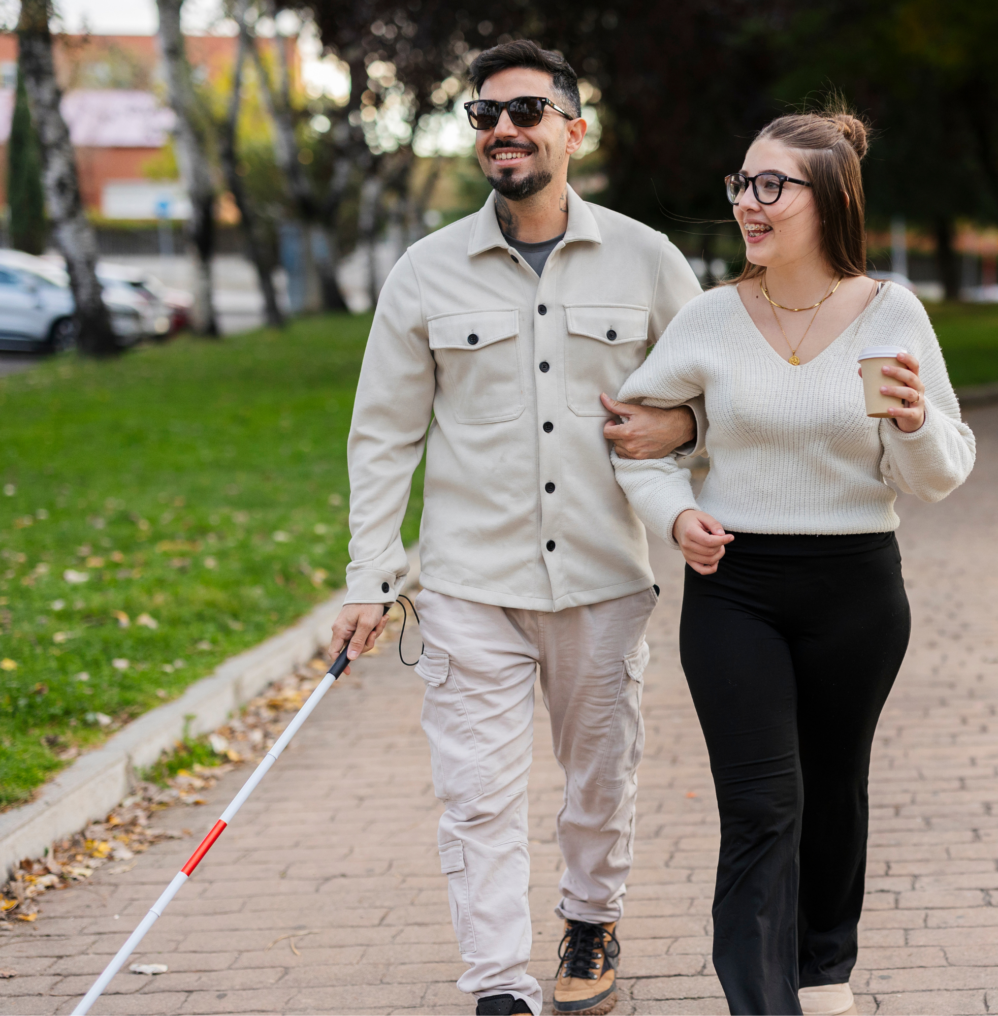 A man using a white cane walks arm in arm with a woman holding a takeaway coffee cup along a tree-lined path.