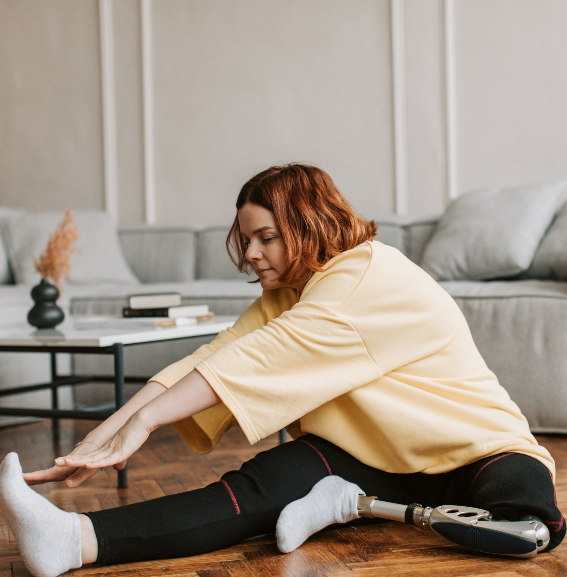 A woman with a prosthetic leg sits on a wooden floor in a living room, stretching forward to touch her foot.