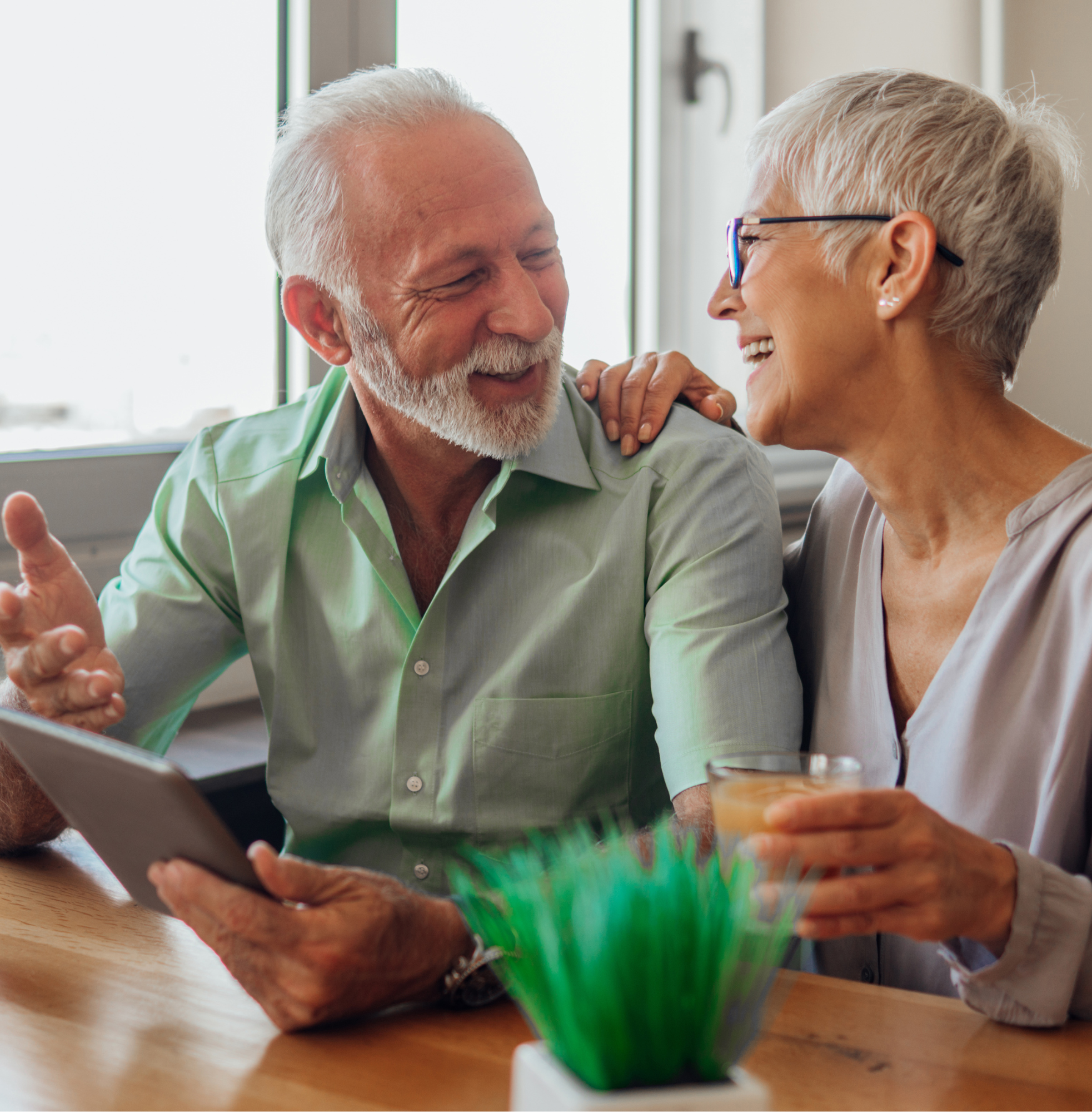 An older couple smiling and talking together at a table while looking at a tablet and holding a drink.