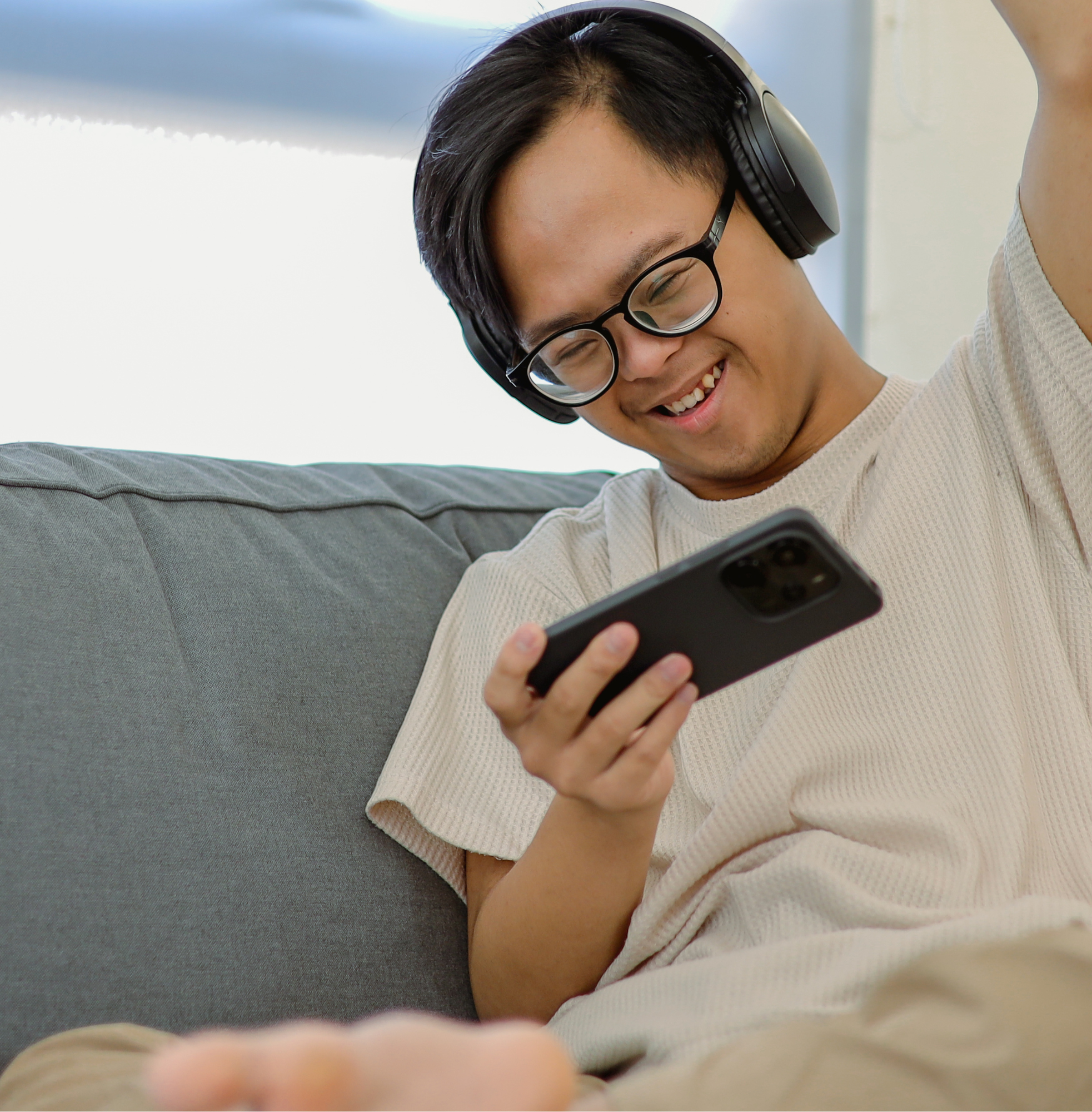A smiling young man with glasses and over-ear headphones sits on a couch, holding a smartphone and raising one arm in celebration.