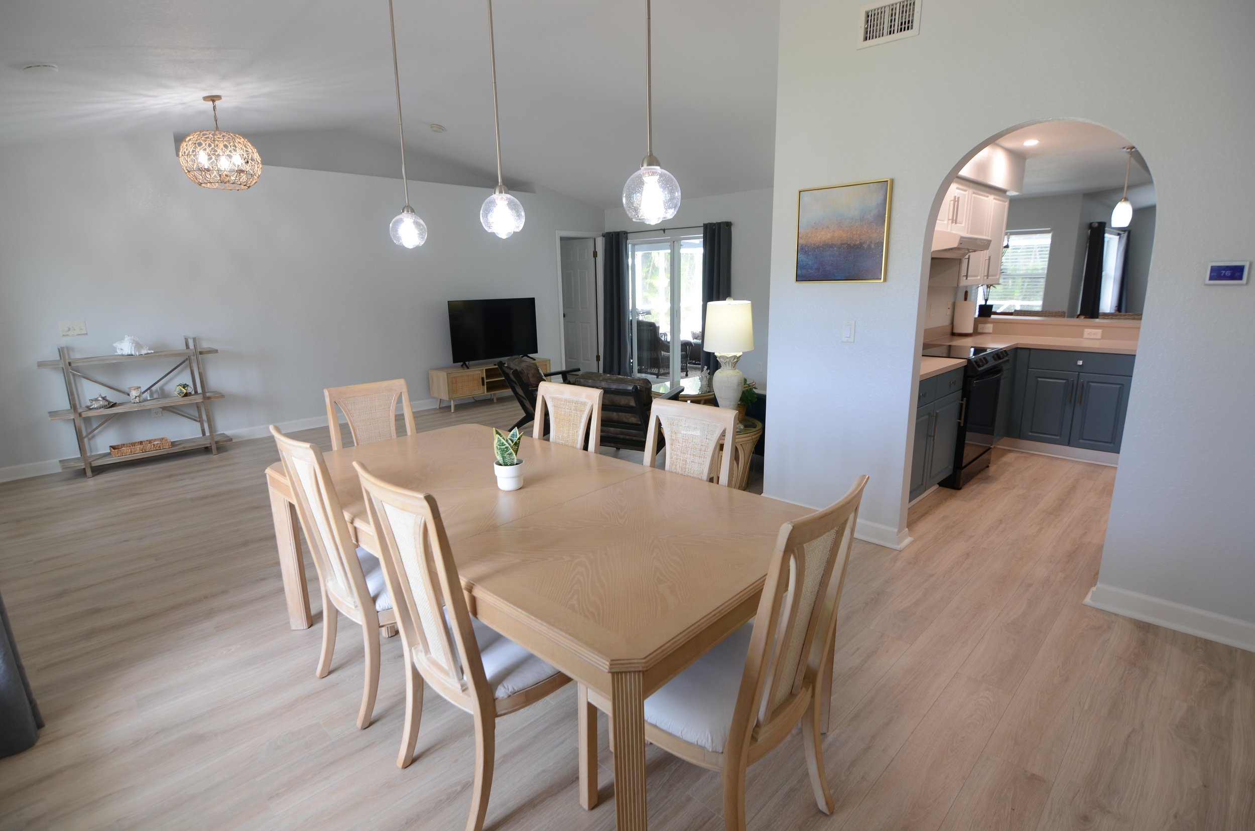 Open concept living and dining area with a wooden dining table, chairs, a sofa, a television, and a kitchen with deep cabinets. Ceiling pendant lights hang above the dining table, and there's a window with dark curtains in the background.