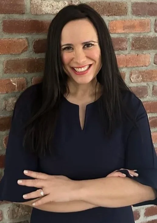 A woman with long dark hair smiling and standing with crossed arms against a brick wall background.
