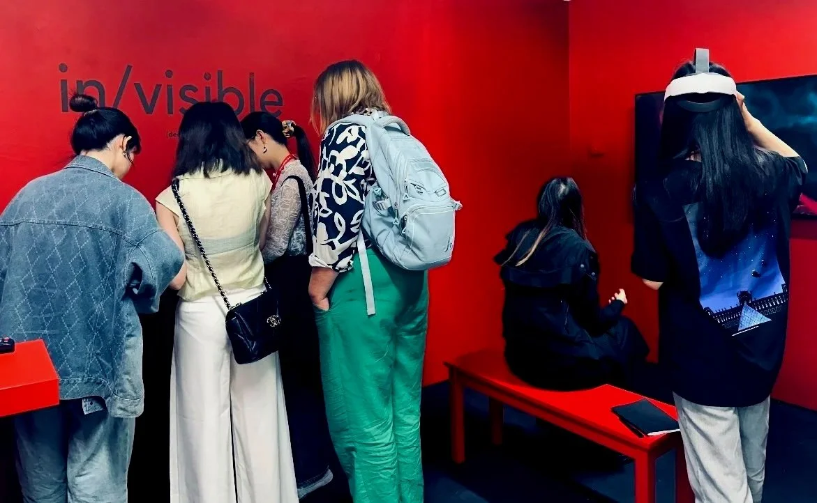 Group of people in an art gallery or museum, observing a display or exhibit on a red wall.