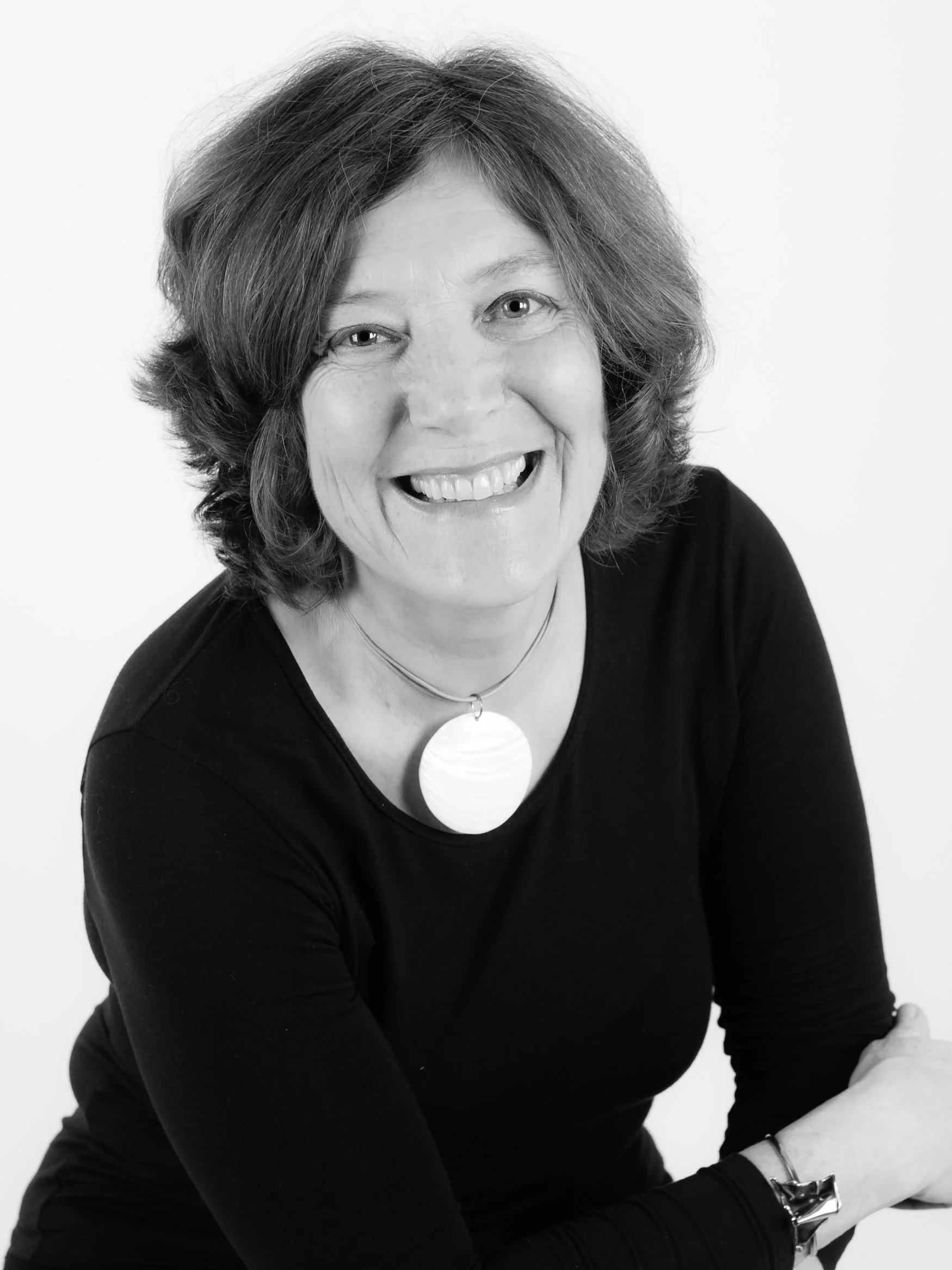 Black and white portrait of a middle-aged woman with short, wavy hair, smiling and looking at the camera, wearing a black top and a large round pendant necklace.