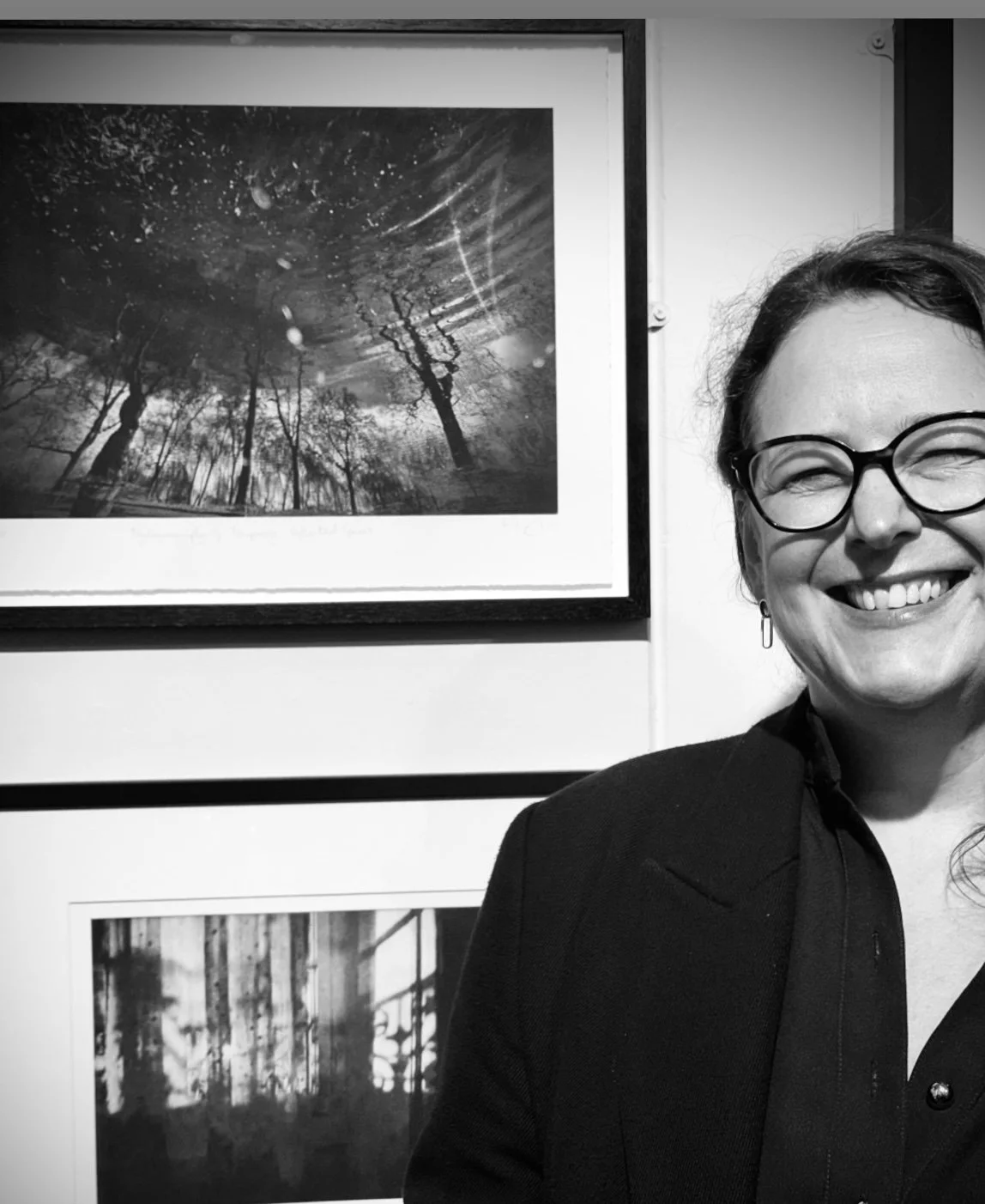 A woman with glasses smiling in front of a framed black and white photograph of trees and a reflected cloudy sky.