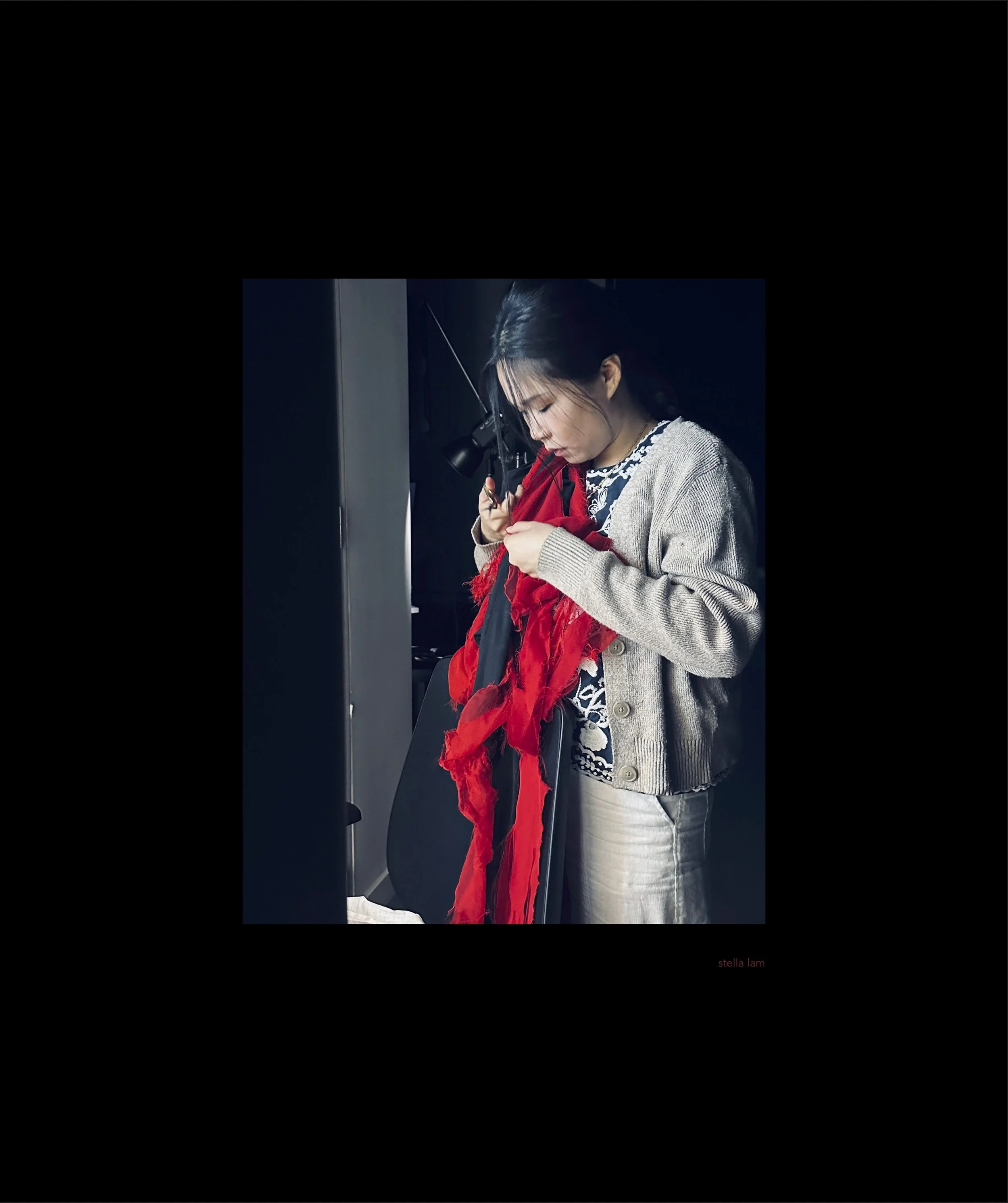 A woman is standing indoors, wearing a light knit cardigan over a patterned shirt. She is holding a red scarf and appears to be focused on arranging or examining it. The background is dark with minimal visible details.