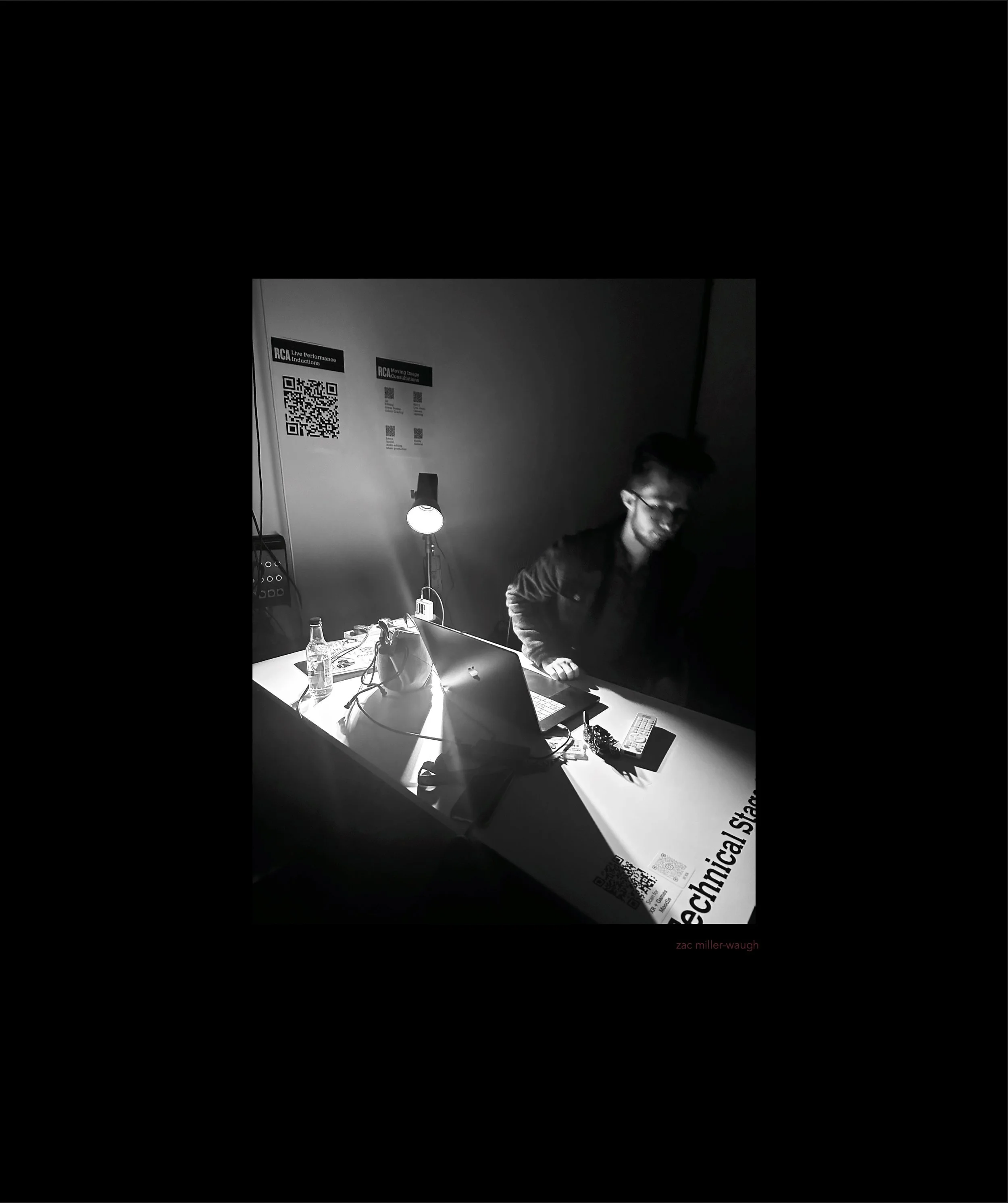 A person wearing glasses and a dark shirt sitting at a desk with a laptop, a water bottle, and electronic devices. There is a lamp illuminating the desk and a white poster on the wall with QR codes and text in the background.