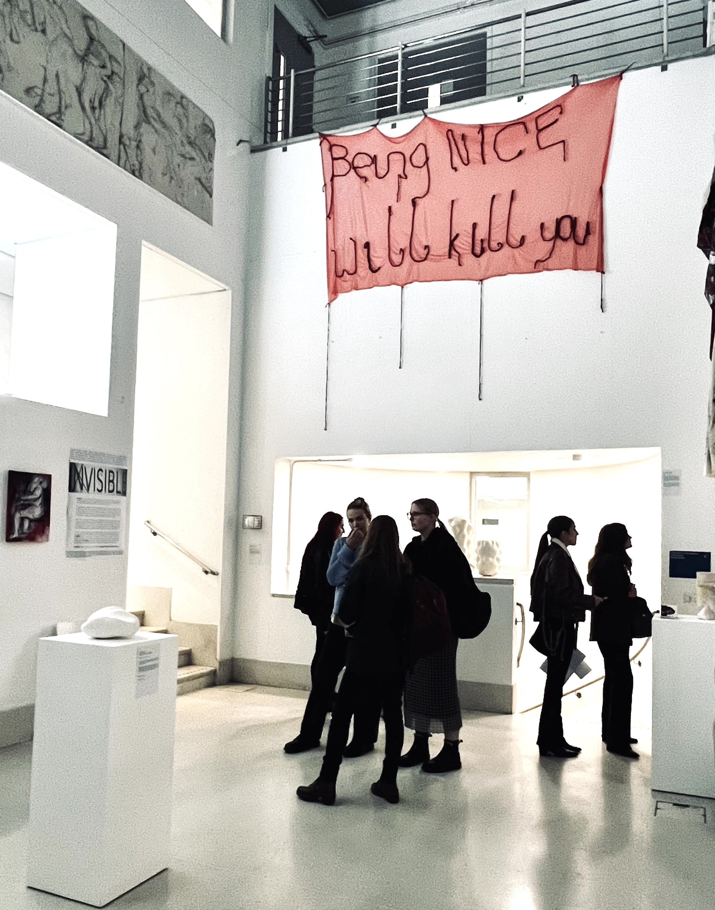 Group of people standing inside a modern art gallery, with some engaged in conversation, in front of white walls displaying artwork and sculptures.