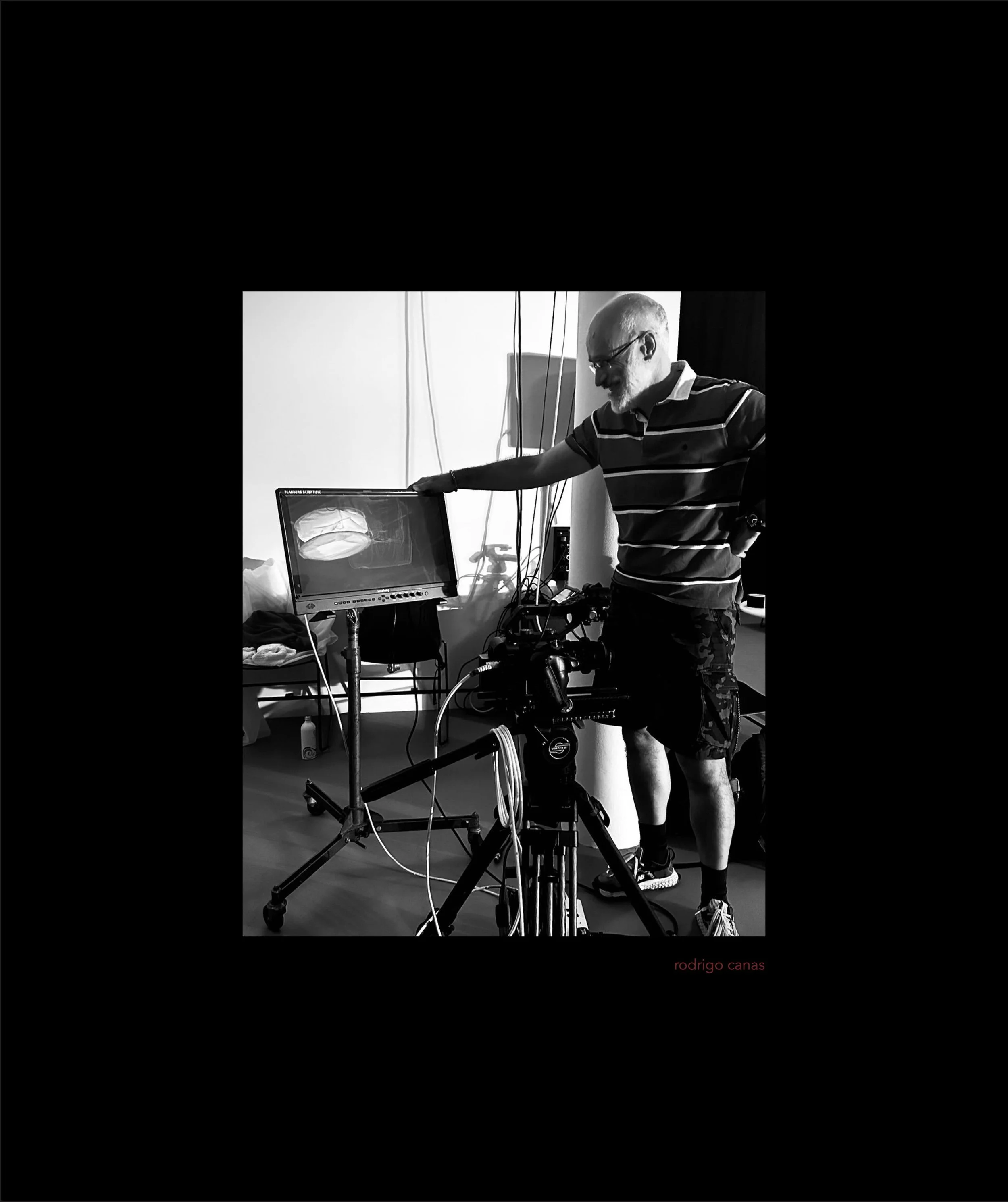 Black and white photo of a man in a striped shirt pointing at a computer screen displaying a 3D brain scan. The man is smiling and appears to be explaining or demonstrating something. The scene is likely in a medical or research setting, with various wires and equipment around. The bottom right corner shows the name 'rodrigo canas' in small red text.