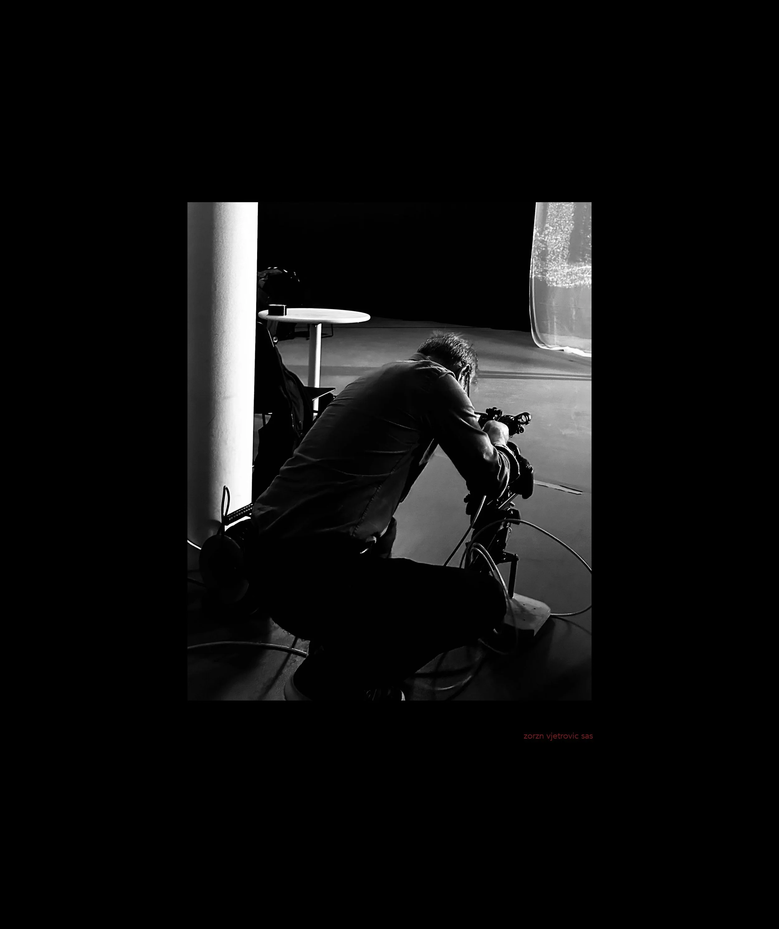 Black and white photo of a man kneeling and adjusting camera equipment in a photography studio, with a table and a large textured reflector or backdrop in the background.