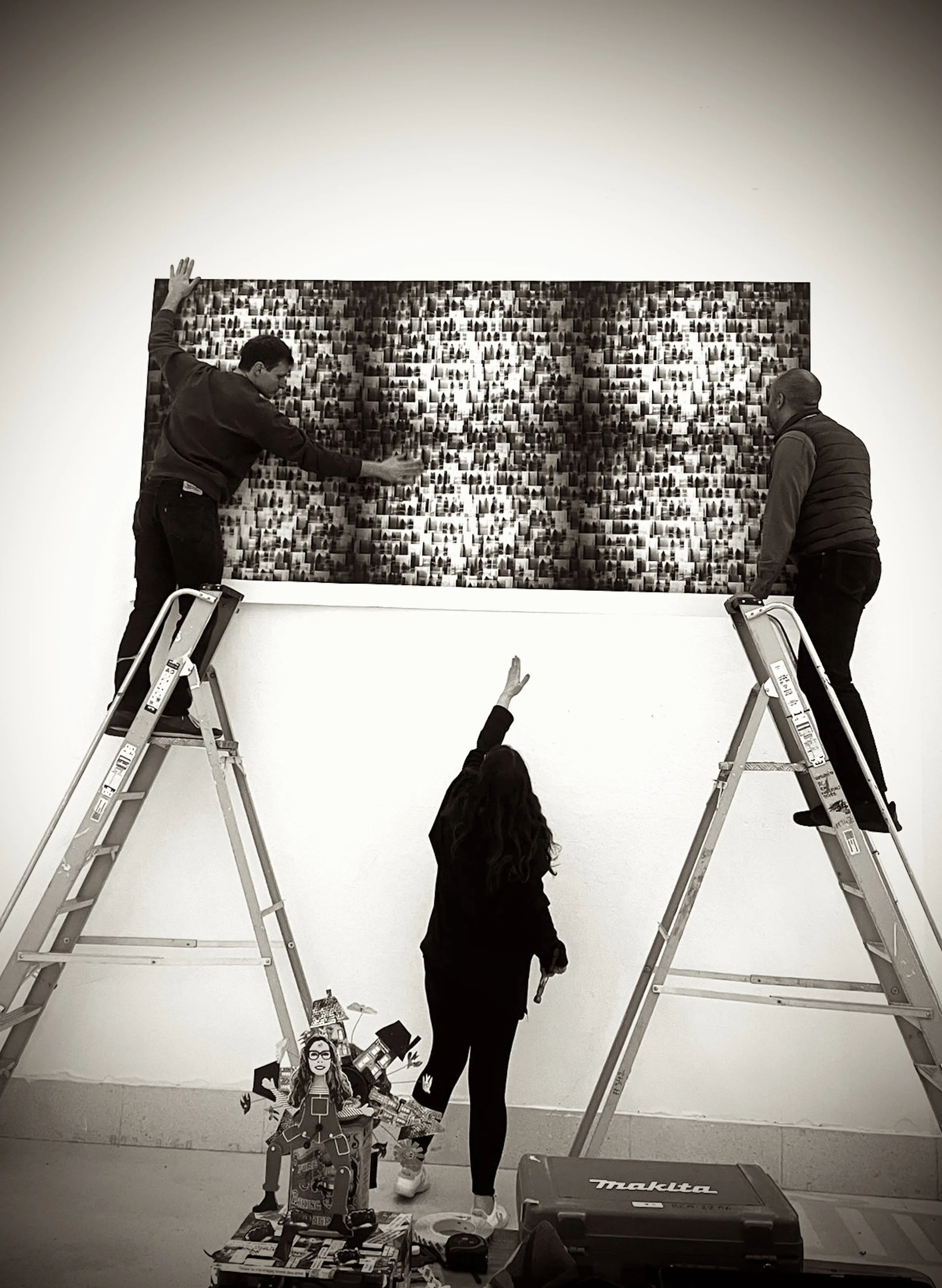People setting up an art display on a white gallery wall with ladders, tools, and artwork on the floor.