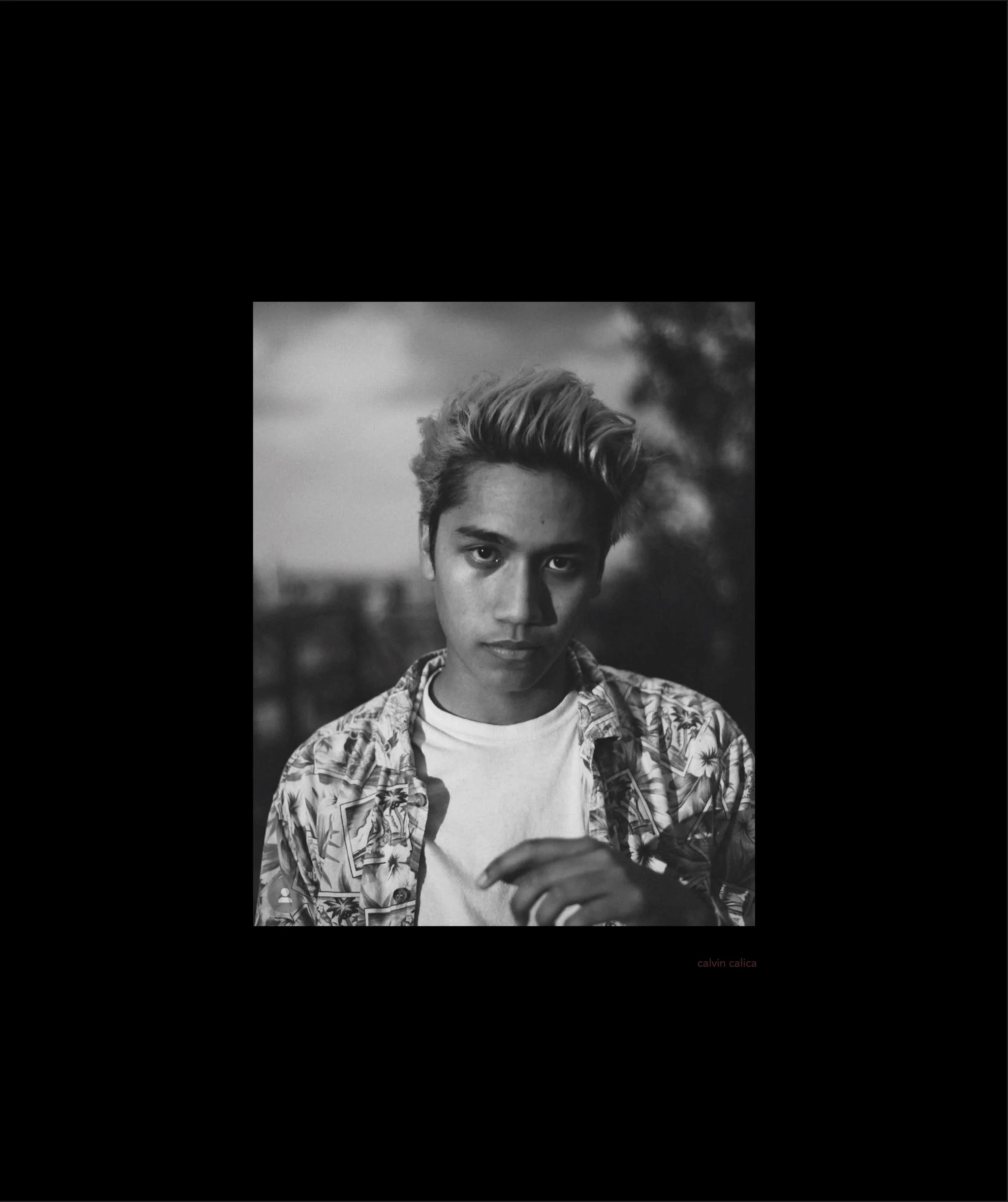 A black and white portrait of a young man with short, styled hair wearing a patterned, button-down shirt over a white t-shirt. The background shows trees and a cloudy sky.