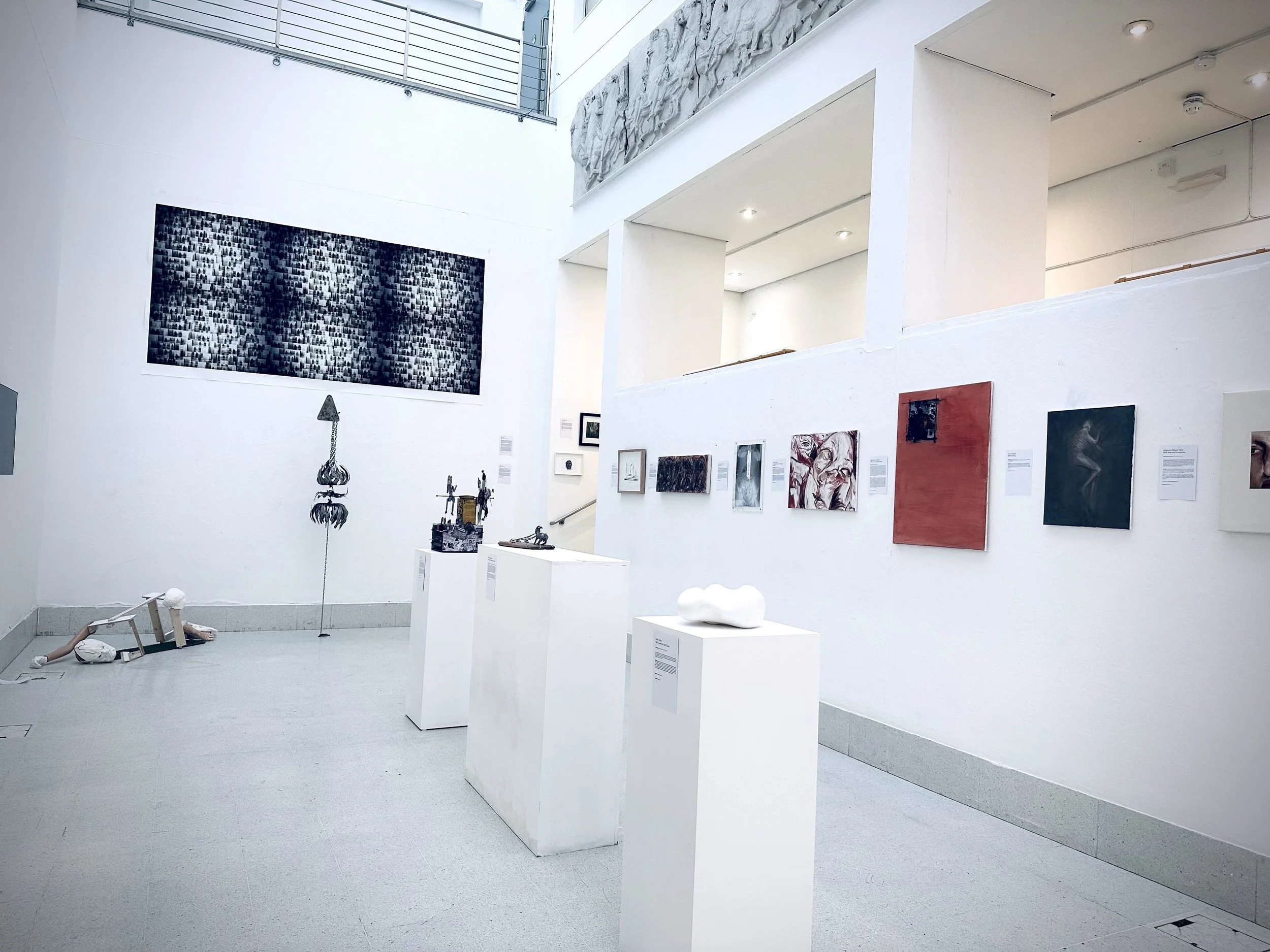 Interior view of an art gallery with white walls, showcasing various artworks, sculptures, and installations. There are ceiling lights illuminating the space, and a staircase leading to an upper level with a metal railing. The gallery features modern and contemporary art pieces.
