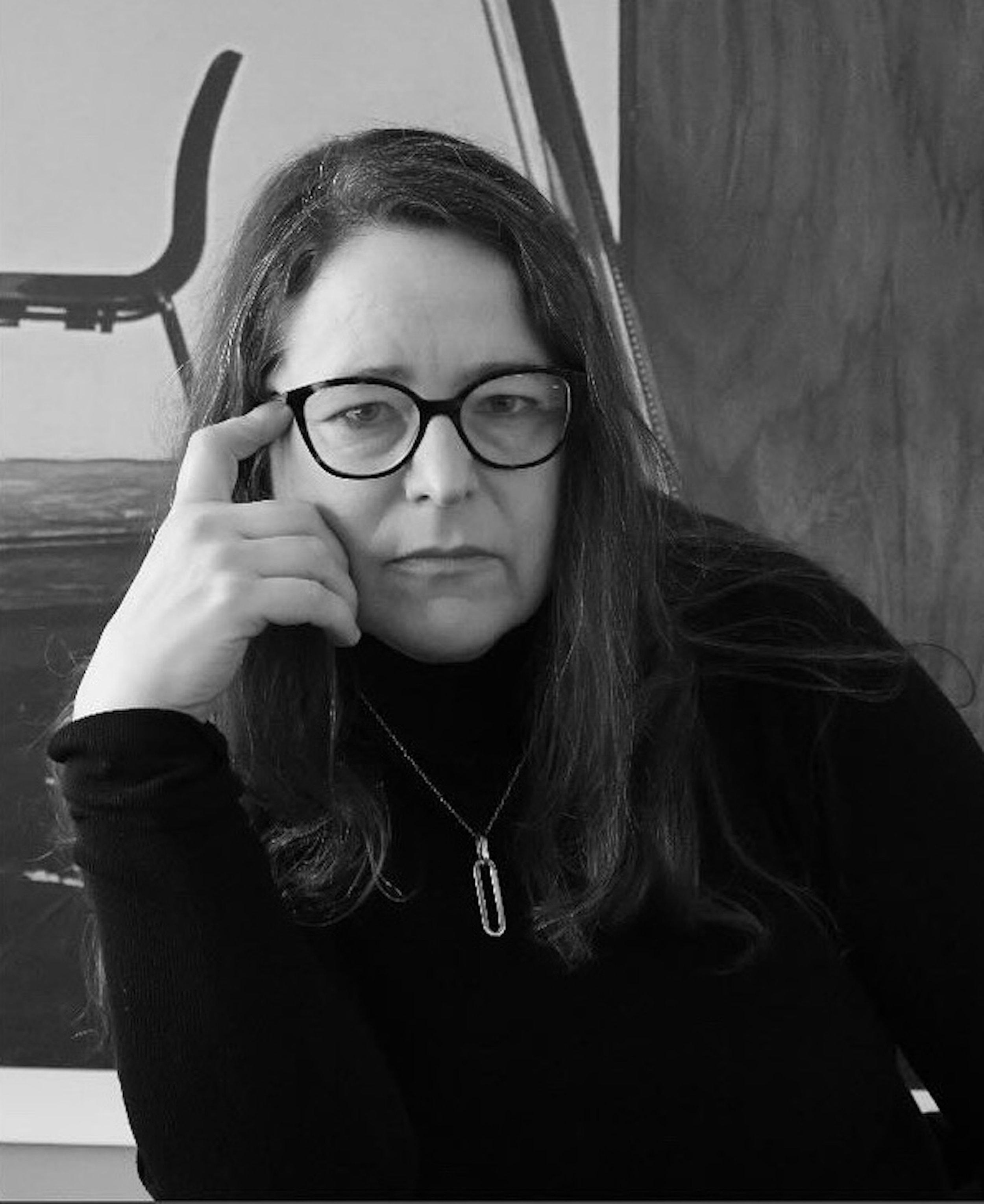 A woman with long hair and glasses, holding her glasses with her right hand, sitting indoors with chairs visible in the background.