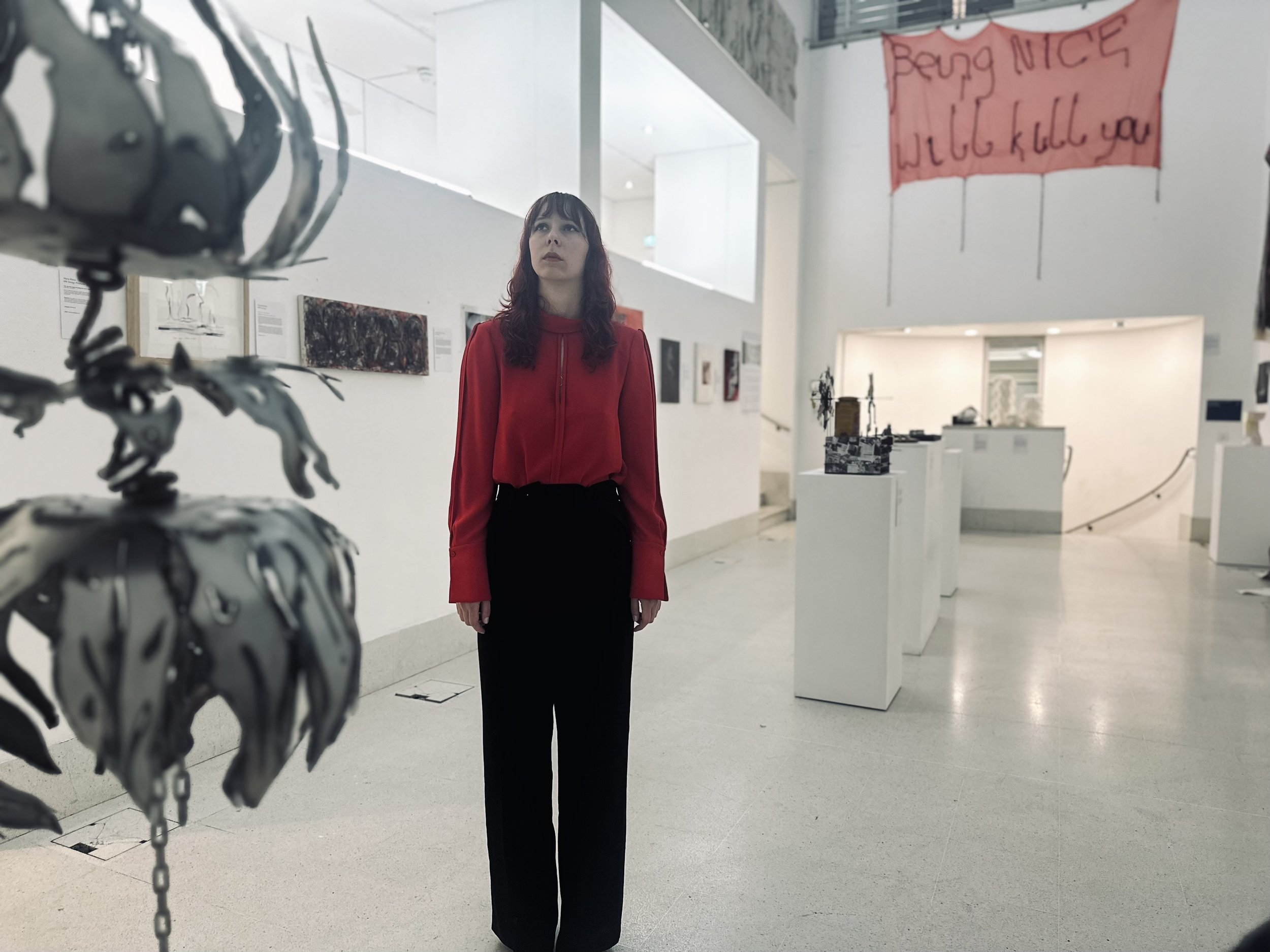A woman with dark hair wearing a red jacket and black pants standing inside an art gallery. The gallery has white walls with artwork and a pink banner hanging that says, "Being Nice Will Kill You." The woman is looking slightly upward with a neutral expression.