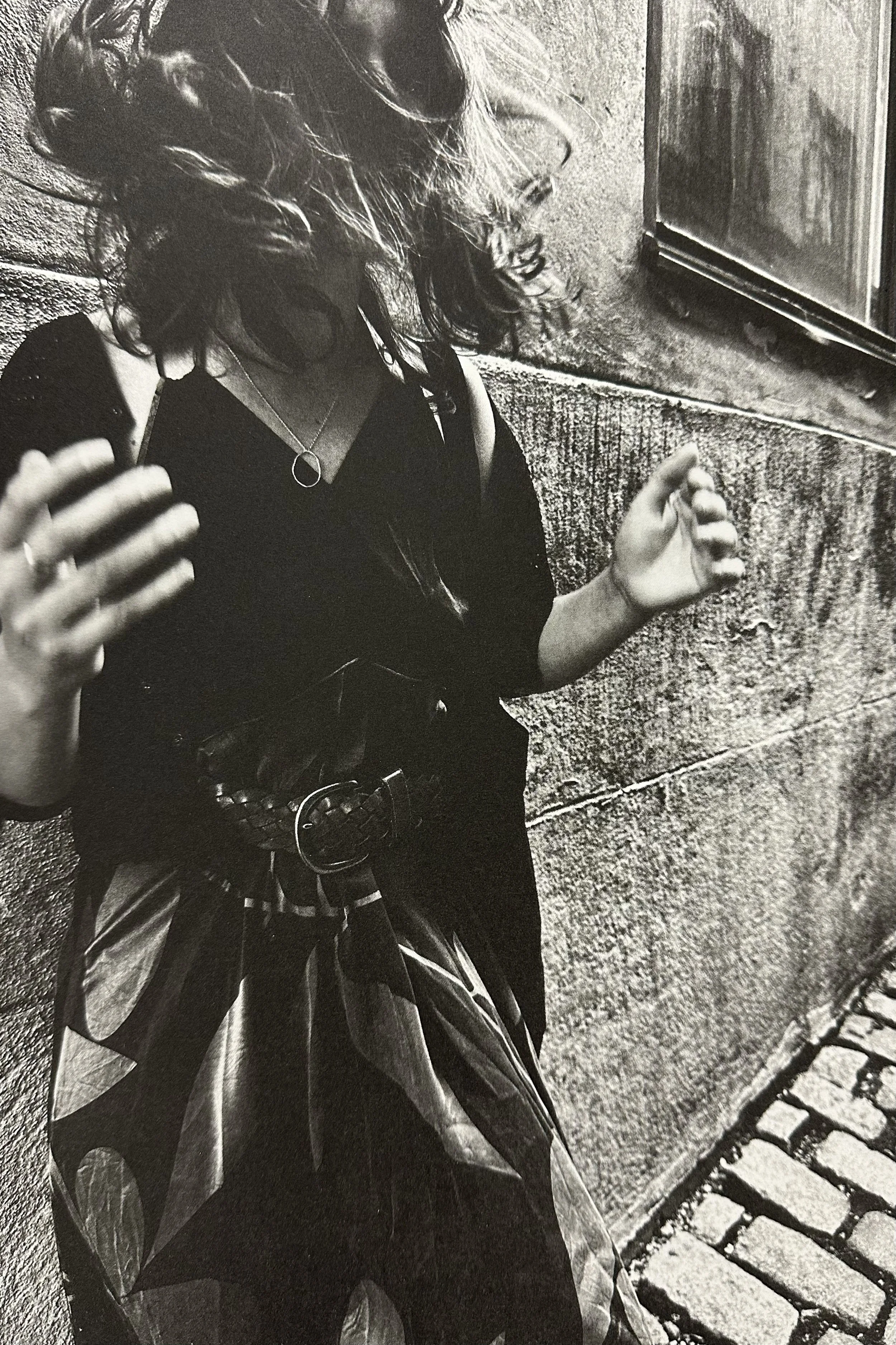 A young woman with curly hair, wearing headphones, a necklace with a circular pendant, and a leather jacket, implies she is listening to music while sitting outdoors near a cobblestone pathway and stone wall.