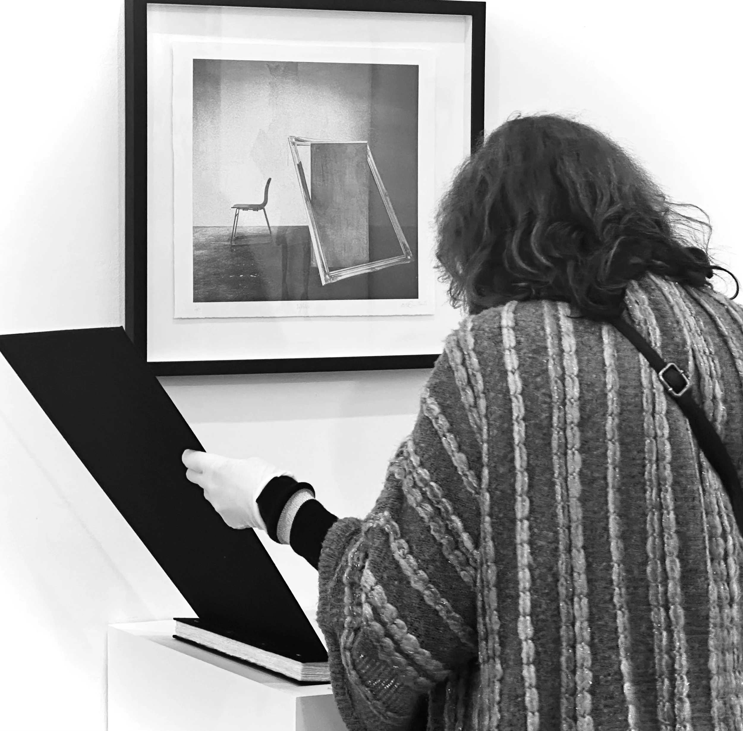 A person with long curly hair is looking at a framed artwork on a wall, which features a photograph of a chair and a mirror in an art gallery or museum.
