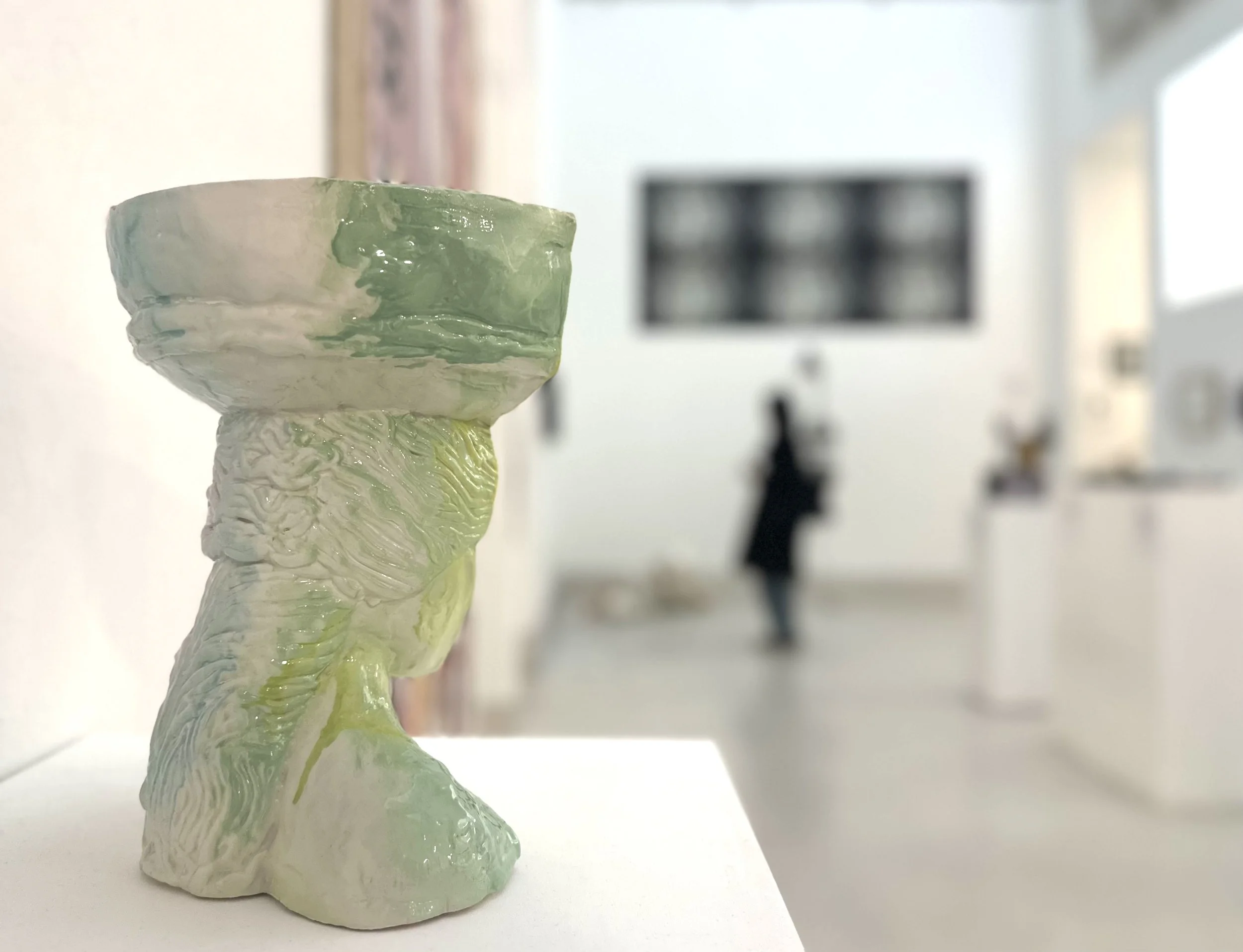 A small abstract ceramic sculpture with green and white glaze, placed on a white surface in a gallery or studio setting with blurred background.