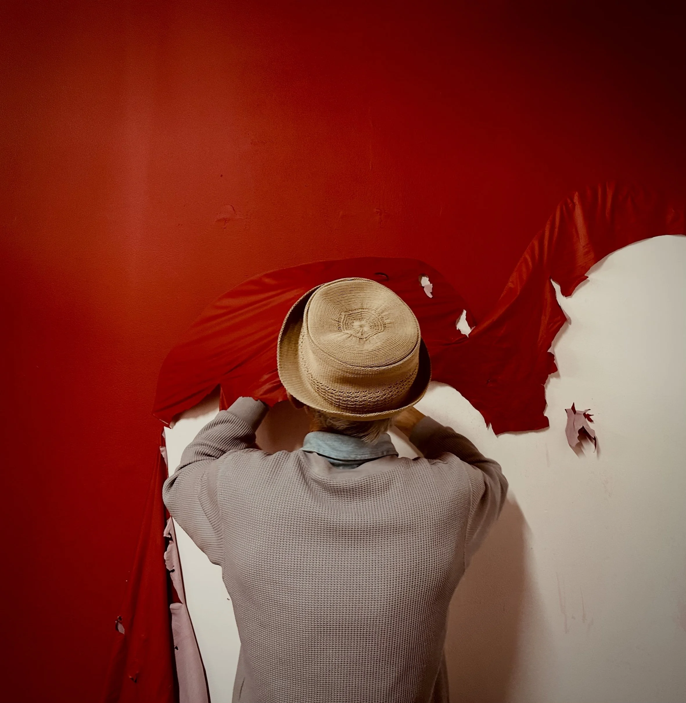 Person in gray long-sleeve shirt with straw hat, partial view, on white floor, red fabric or curtain in the background.