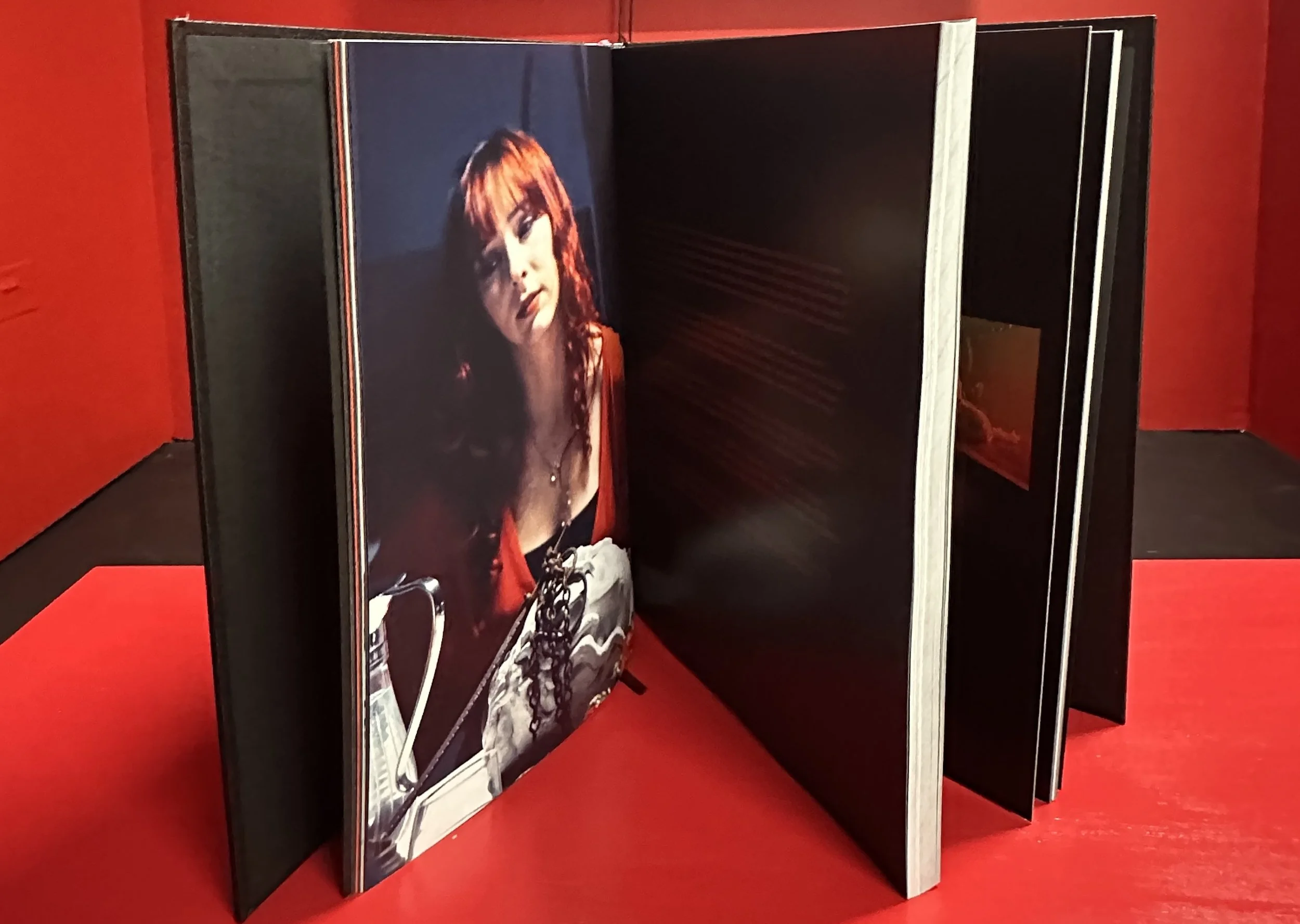 A large open photo album or scrapbook with a picture of a woman with red hair, wearing a black outfit, and holding a camera, displayed on the left page, set against a red background.