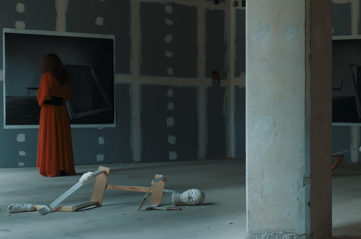 An art installation featuring a seated woman in a long red dress, a distorted sculpture on the floor, and framed photographs on unfinished walls in a gallery space.