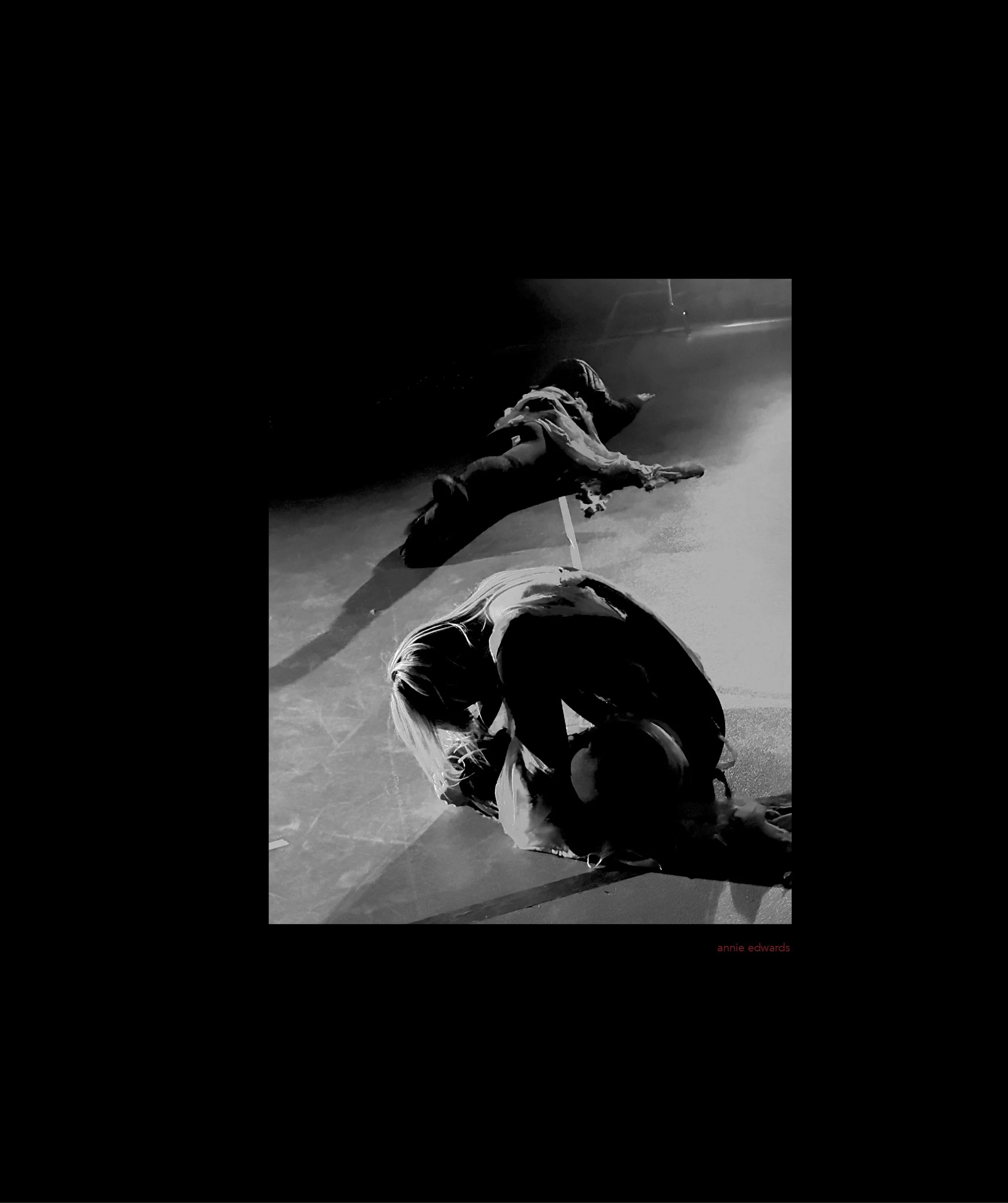 A person lying face down on a dark road at night, with another person crouched nearby. The scene is illuminated by a single light source, casting shadows. The text 'annie edwards' is visible in red at the bottom right corner of the image.
