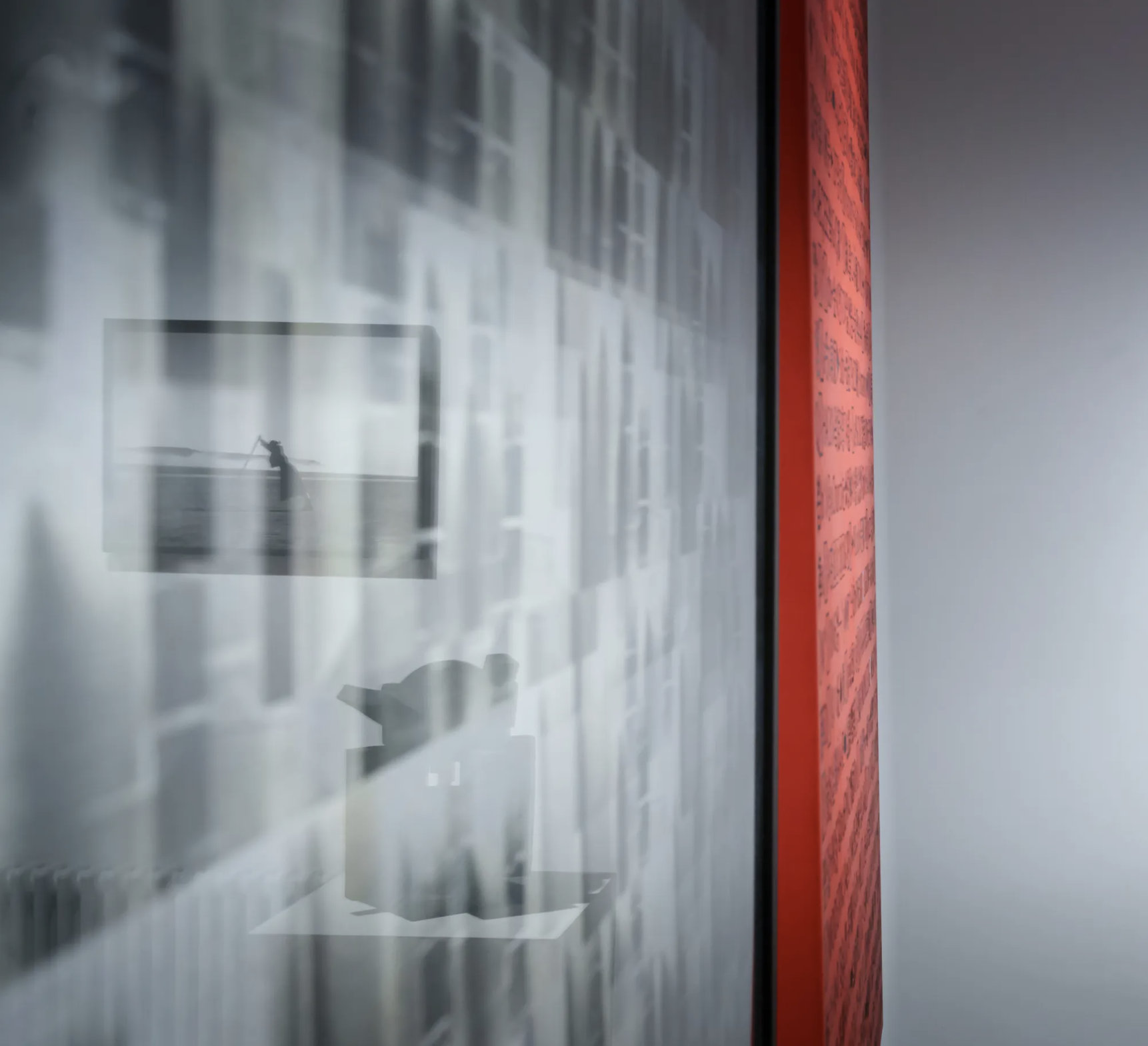 Reflected image of a person playing a guitar, seen through a glass wall with various pictures or posters. The scene appears to be indoors, possibly in a gallery or studio, with a red strip or artwork on one side.