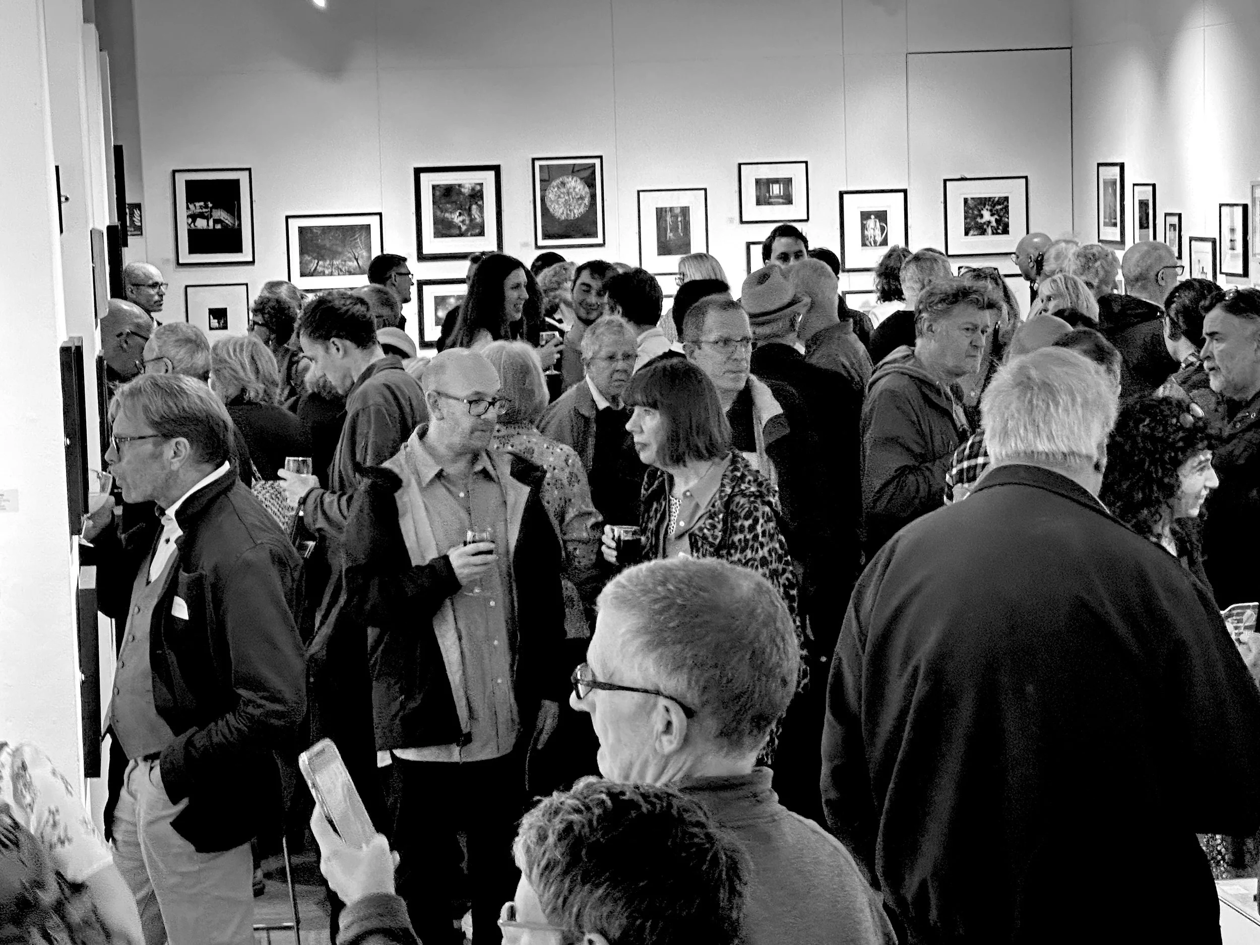 A crowded art gallery with numerous framed photographs hanging on the walls and many people engaged in conversation and viewing the artwork.
