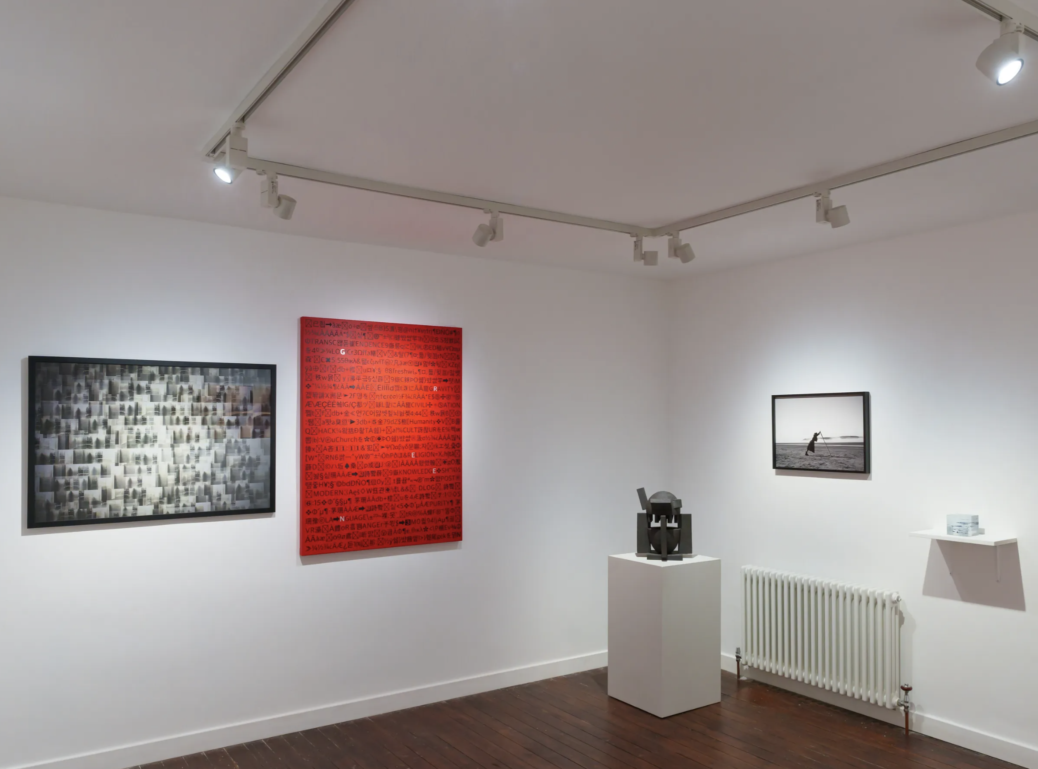 An art gallery with several framed artwork pieces on the white walls, including a black and white abstract art piece, a red artwork with text, and a black-and-white photograph of a person on the beach. There are two white pedestals with sculptures and a small white table with books nearby. Track lighting illuminates the artwork.