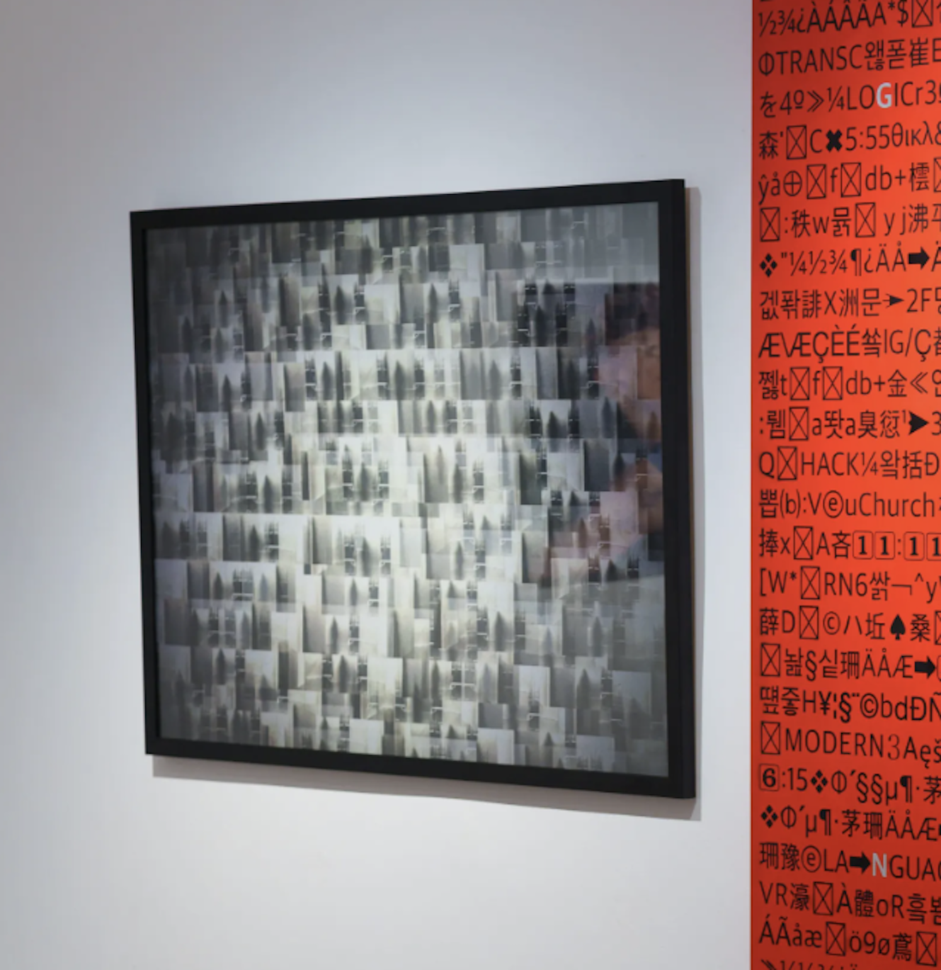 Abstract pixelated artwork with a black frame, hanging on a white wall to the left of a red panel with various symbols, text, and characters.