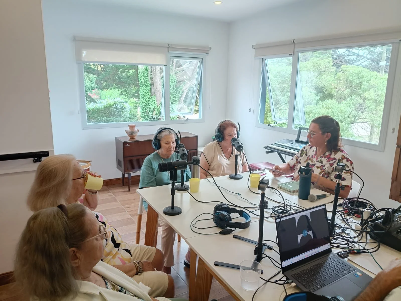Cinco mujeres participando en una grabación de podcast en una habitación con ventanas grandes y vistas verdes, con equipos de grabación y computadoras en la mesa.