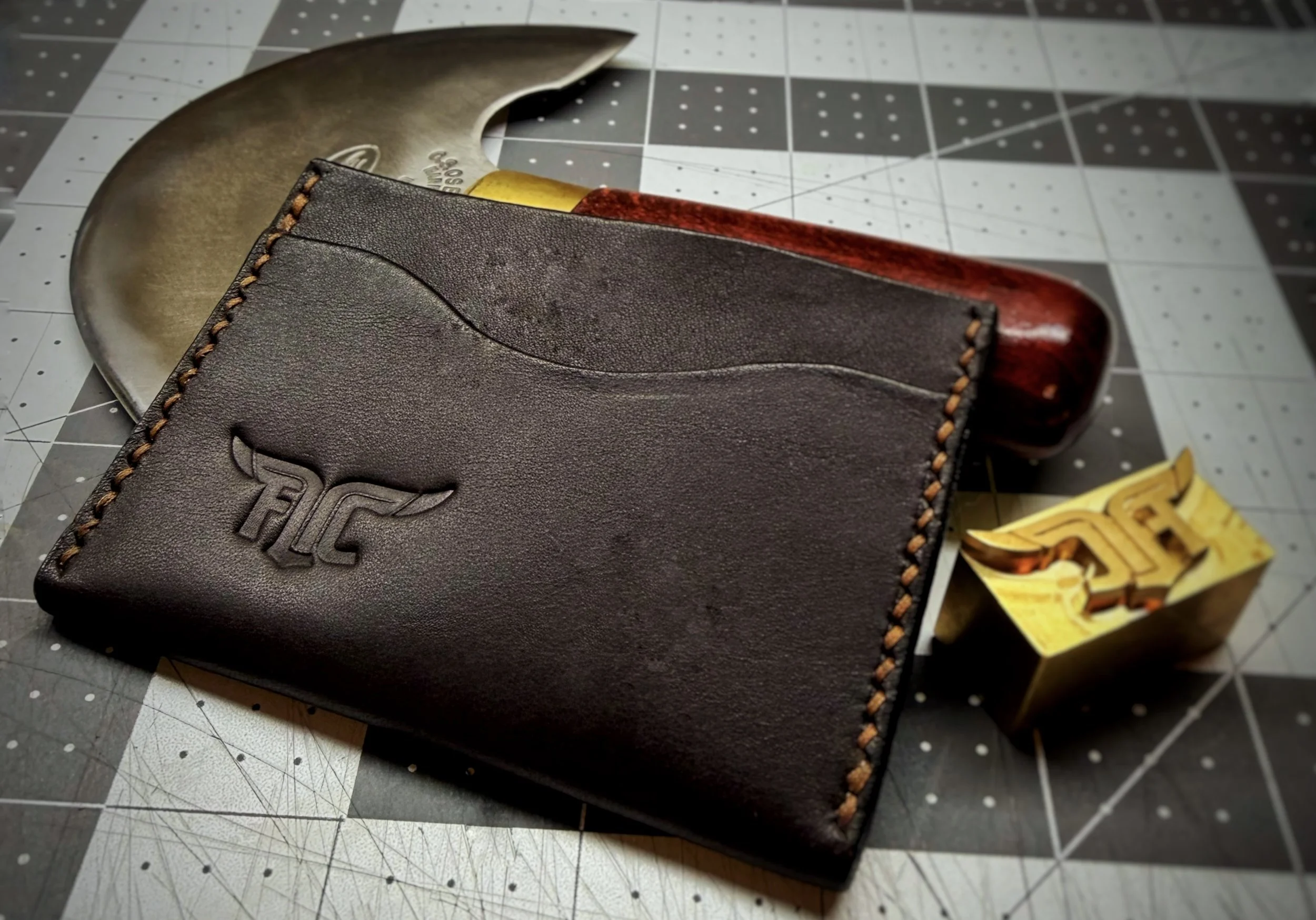 A black leather wallet with orange stitching placed on a cutting mat, containing a folding pocket knife with a wooden handle, a gold-colored pocket clip, and a gold-colored item, possibly a small tool or accessory.