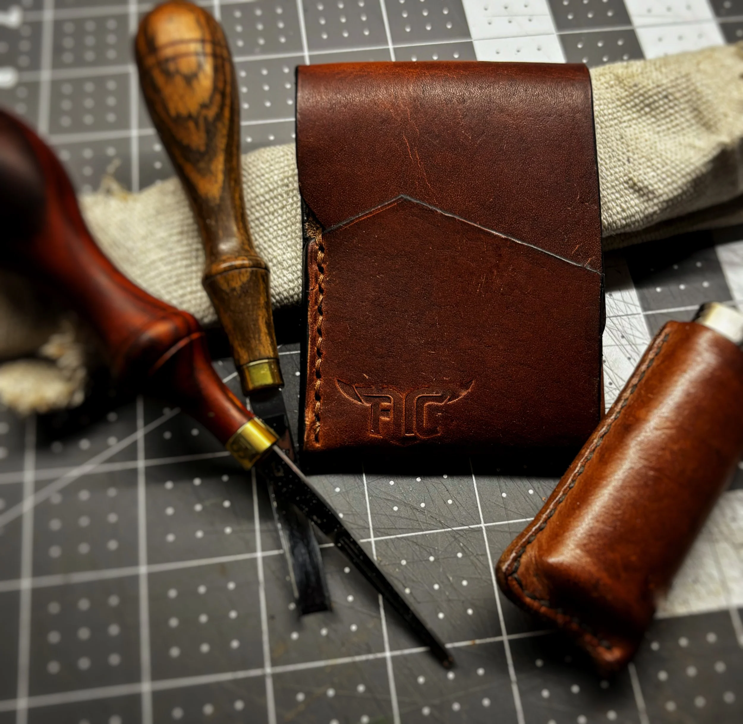 Close-up of a leather wallet, two carving chisels with wooden handles, and a leather tool roll, all placed on a cutting mat with a grid pattern.
