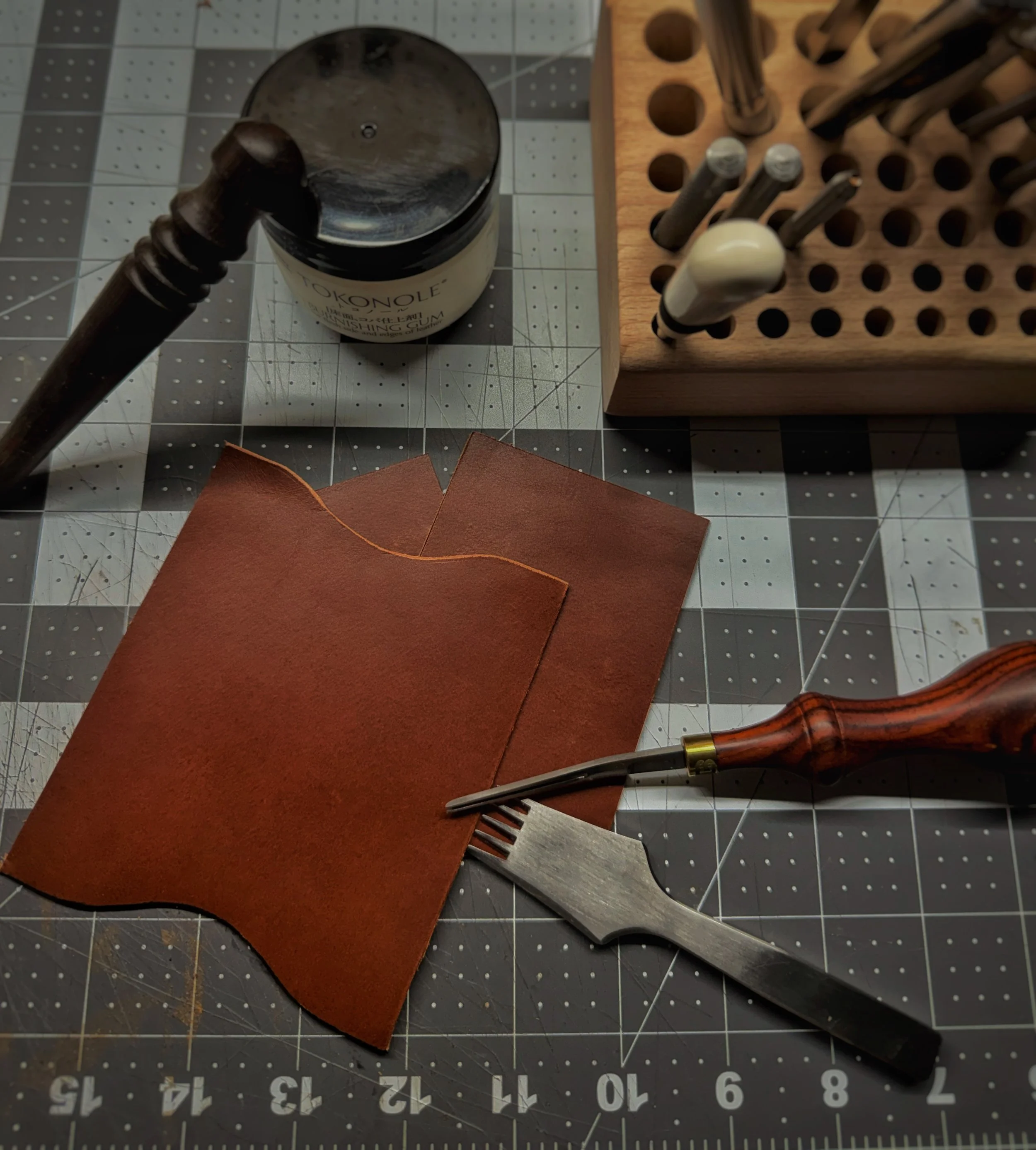 Leatherworking tools including a mallet, a piece of reddish-brown leather, a metal and wooden pricking iron, and a jar of gum on a cutting mat with measuring grid.