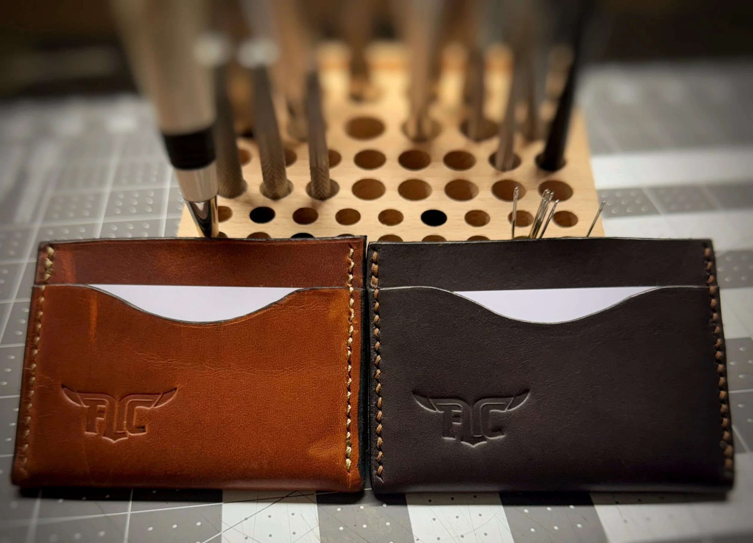 Two leather wallets, one brown and one black, with a logo, placed on a cutting mat. In the background, there is a wooden holder with various metal tools.