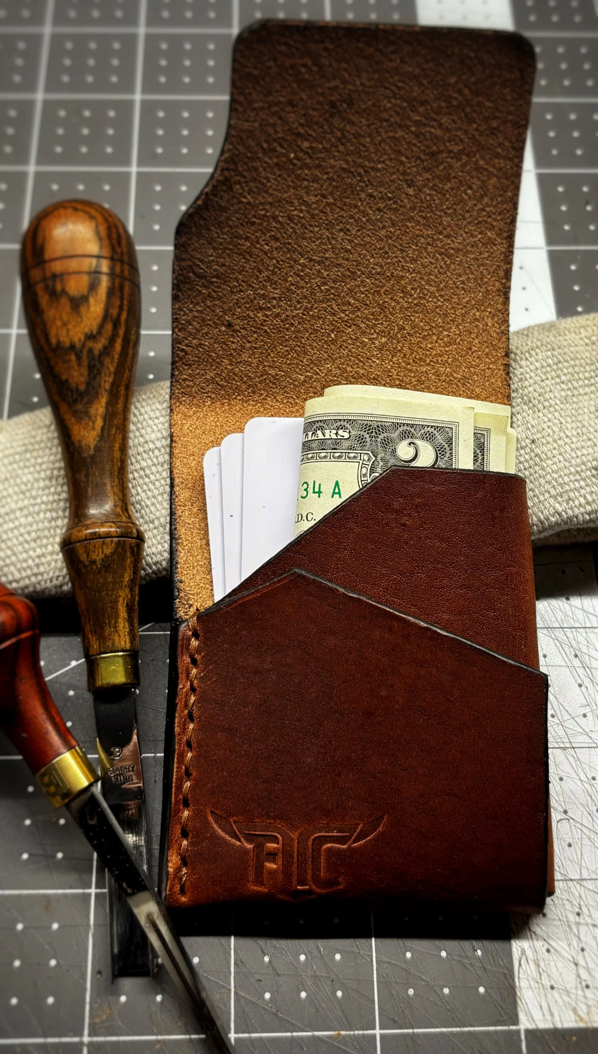 Brown leather wallet with cash sticking out, a leather strop, and carving tools on a cutting mat.
