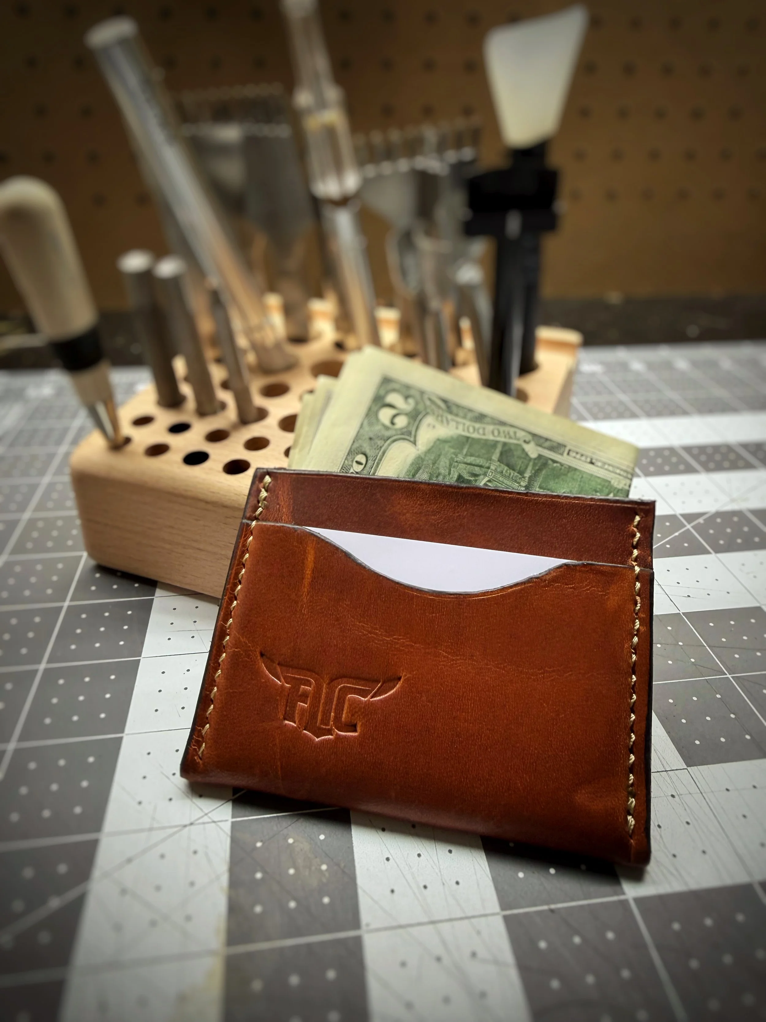 A leather wallet with embossed logo, containing a few dollar bills, on a craft cutting mat with a tool holder in the background.