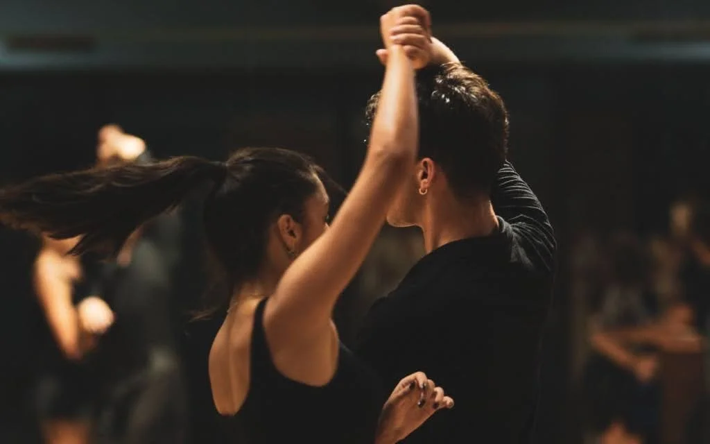 Cosa indossare a lezione di ballo: una coppia balla in sala con abiti neri comodi, lei con un top senza maniche e la coda di cavallo in movimento, lui con una maglia nera a maniche lunghe