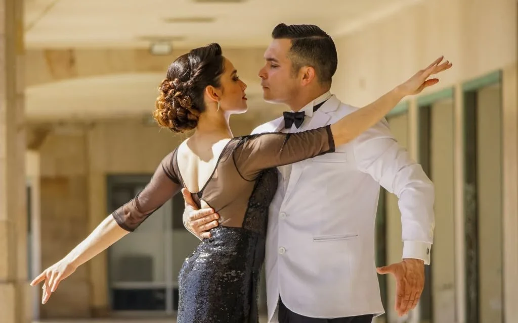 Ballare in coppia: un uomo in giacca bianca e papillon e una donna con abito nero a paillettes e capelli raccolti ballano in posizione di abbraccio sotto un portico