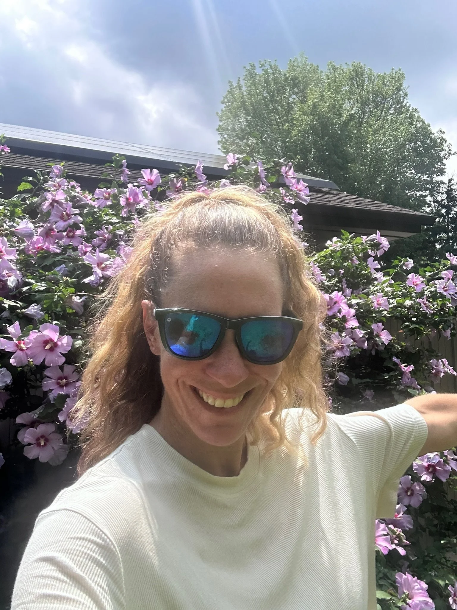 A smiling woman with curly blonde hair and sunglasses taking a selfie outdoors in front of purple flowers, with a house roof and a large green tree in the background under a partly cloudy sky.