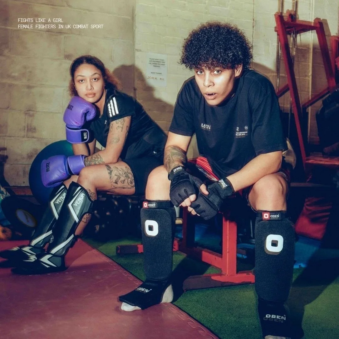 Two female fighters in boxing gloves and combat gear, sitting in a gym. One is wearing purple gloves, and the other is in black gear with shin guards. They appear ready for a fight.