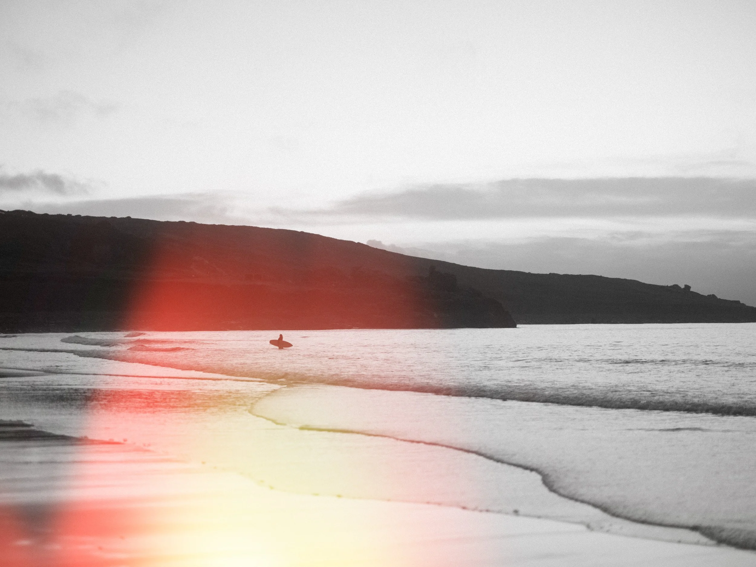 Silhouette of a person with a surfboard walking along the shoreline at sunset, with hills and a cloudy sky in the background.
