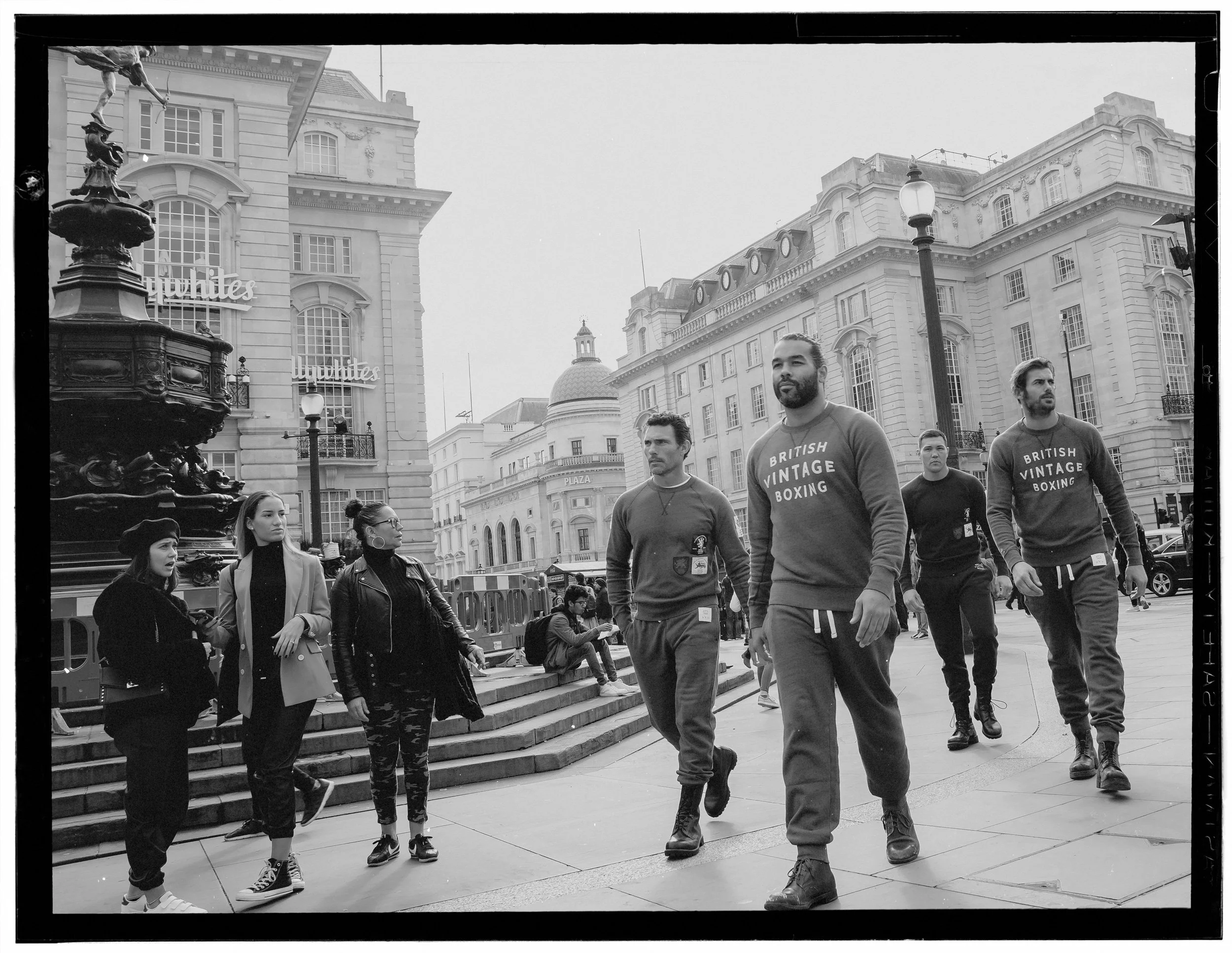A group of people walking on steps in an urban area with historic style buildings in the background, including a fountain and a sign that reads 'Dainties'.