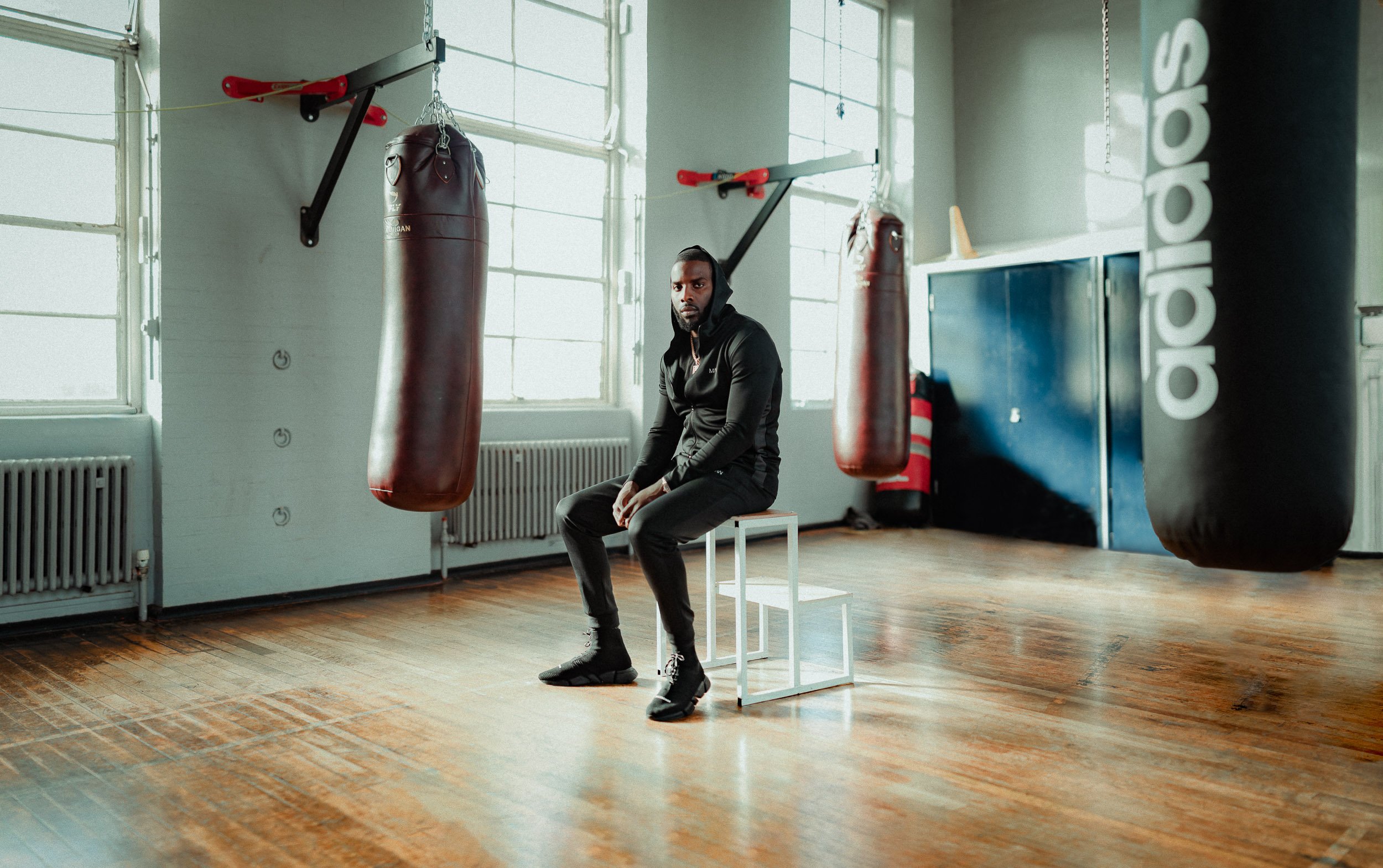 A man in black athletic clothing sits on a white stool in a boxing gym, looking toward the camera. There are two red punching bags hanging from the ceiling, and a large black punching bag with some text on it in the background. The gym has large windows letting in natural light and wooden floors.