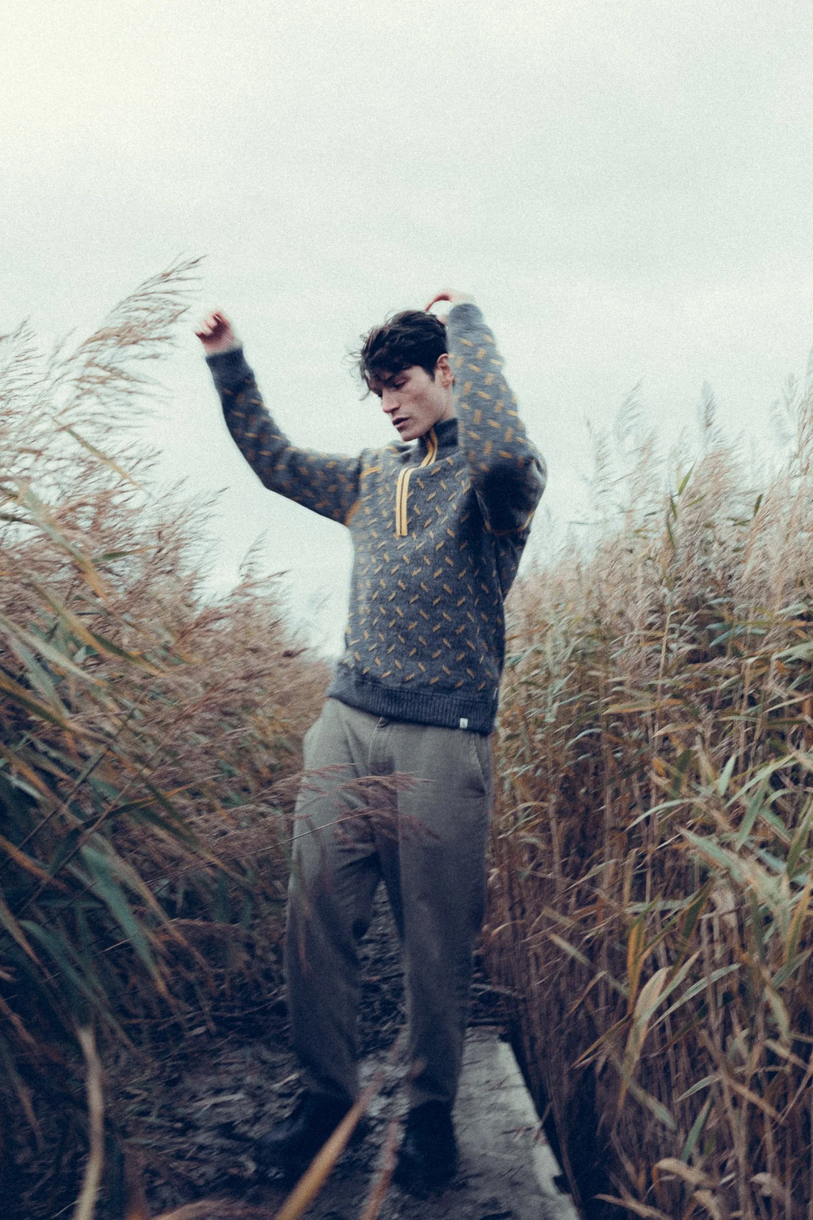A young man with dark hair standing in a narrow waterway surrounded by tall, brownish-green reeds, with his arms raised and eyes closed, dressed in a patterned jacket and gray pants.