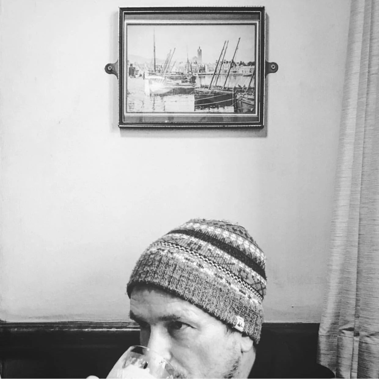 A man wearing a knit cap is drinking from a glass, with a framed print of boats on water hanging on the wall behind him.