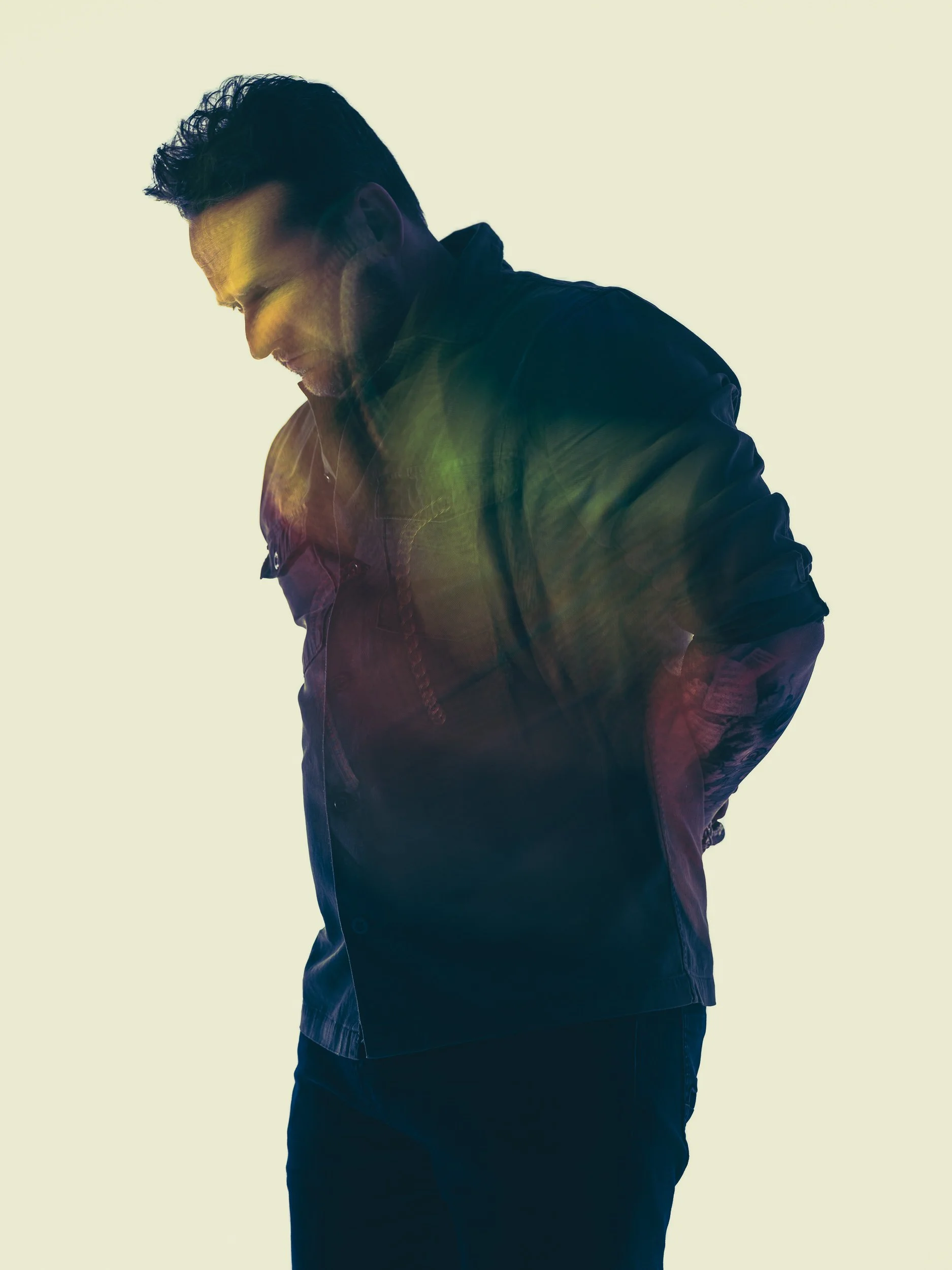 Side profile of a man wearing a black jacket, looking down with a thoughtful expression, against a plain light background.